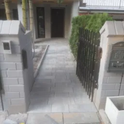 Ścieżka z szarych, prostokątnych kostek prowadząca do drzwi wejściowych domu, ograniczona z jednej strony metalową furtką i zielonymi krzewami, a z drugiej słupkami z cegieł z domofonem i skrzynką...