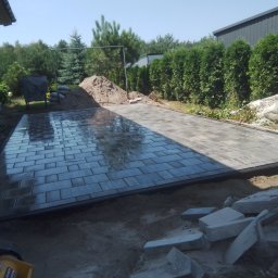 Malbruk - Układanie kostki brukowej w trakcie prac: widoczna część ułożonej, mokrej kostki, hałda piasku, worki z kruszywem i narzędzia na placu budowy w słoneczny dzień.