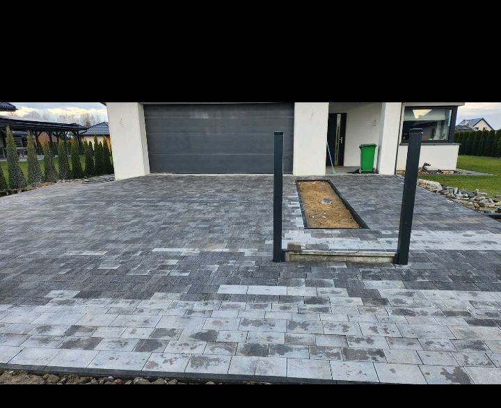 Ułożona szara kostka brukowa na podjeździe przed domem z widocznym miejscem na trawnik, czarne słupki pergoli i zamkniętą bramą garażową.