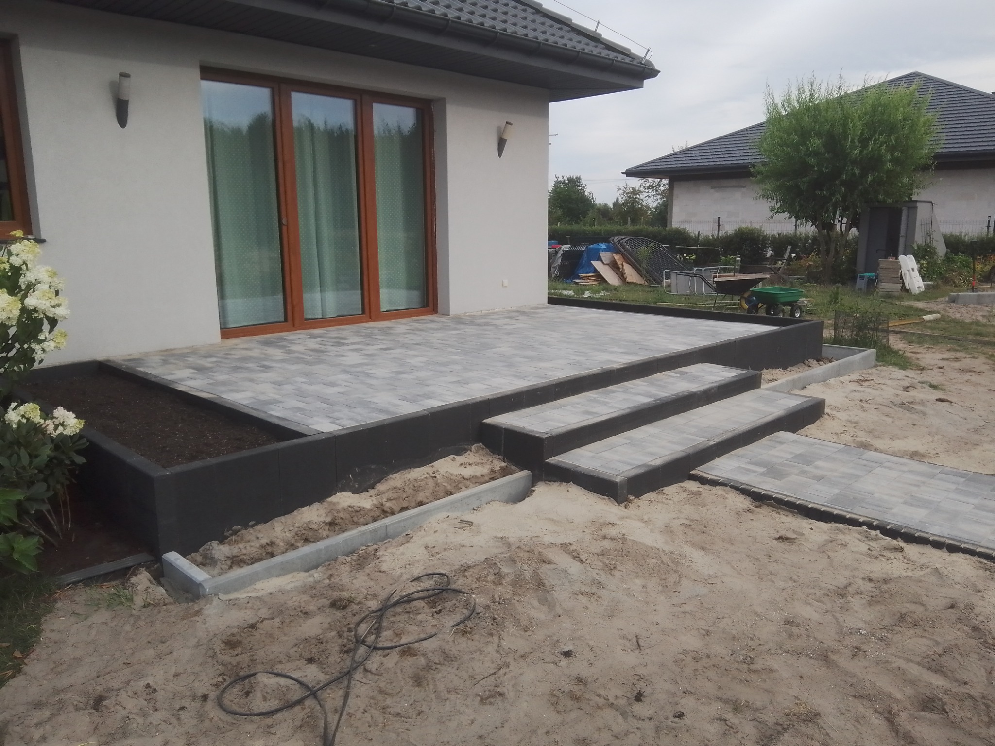 Wykończony taras z szarej kostki brukowej, z trzema schodkami i rabatą na kwiaty, widok na dom i ogród w tle, teren wokół w trakcie prac ziemnych.
