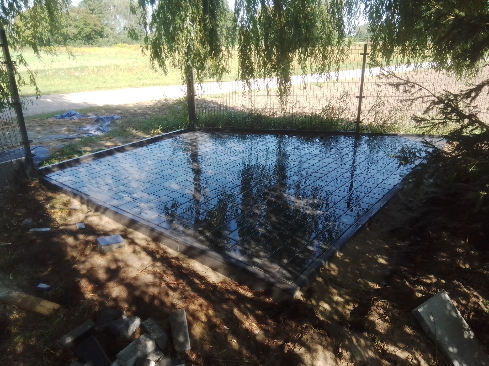 Świeżo ułożona, mokra, ciemna kostka brukowa tworząca kwadratowy taras, otoczona szarą obwódką; w tle widoczna trawa, drzewa i siatka ogrodzeniowa.