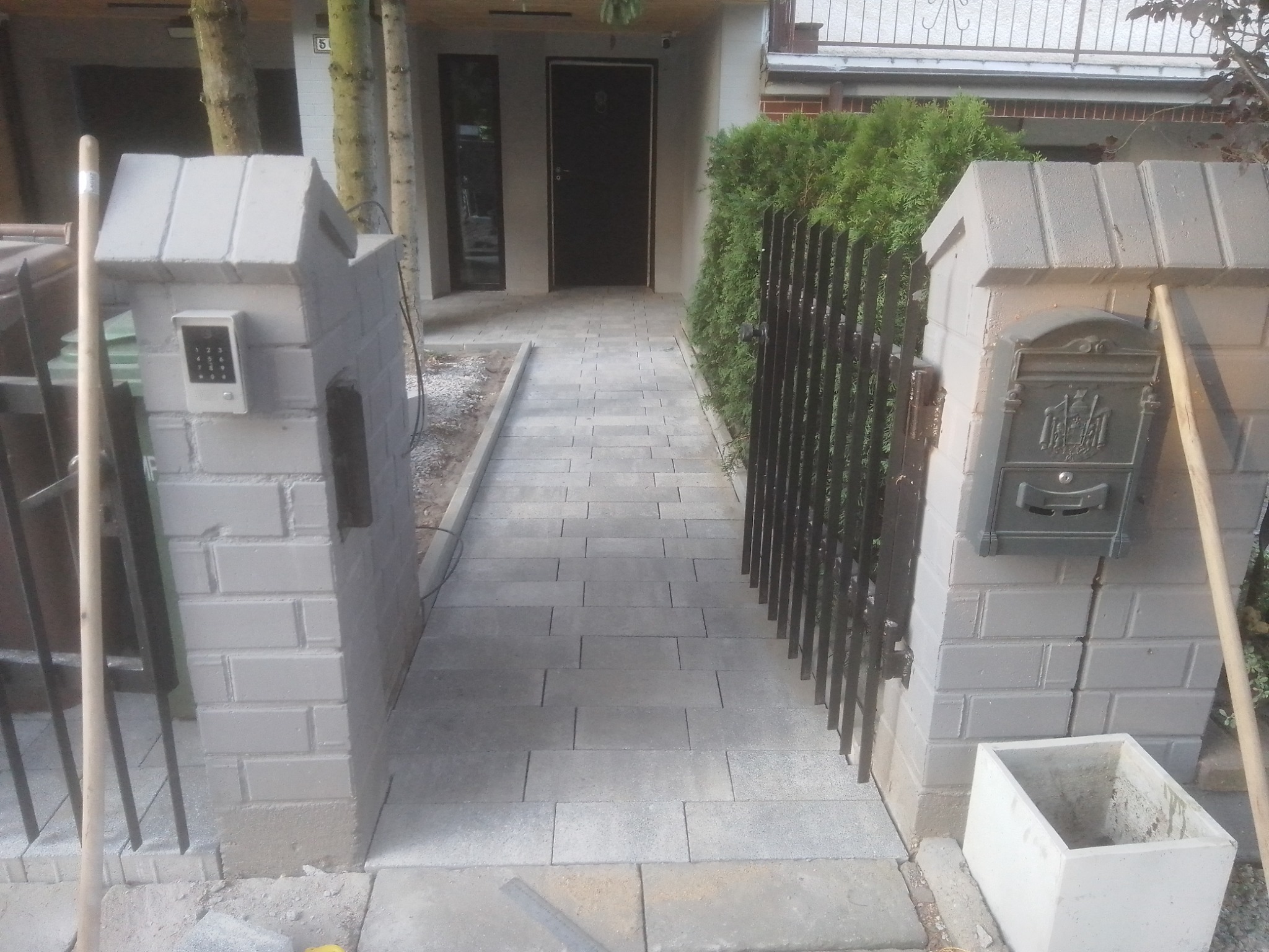 Ścieżka z szarych, prostokątnych kostek prowadząca do drzwi wejściowych domu, ograniczona z jednej strony metalową furtką i zielonymi krzewami, a z drugiej słupkami z cegieł z domofonem i skrzynką...