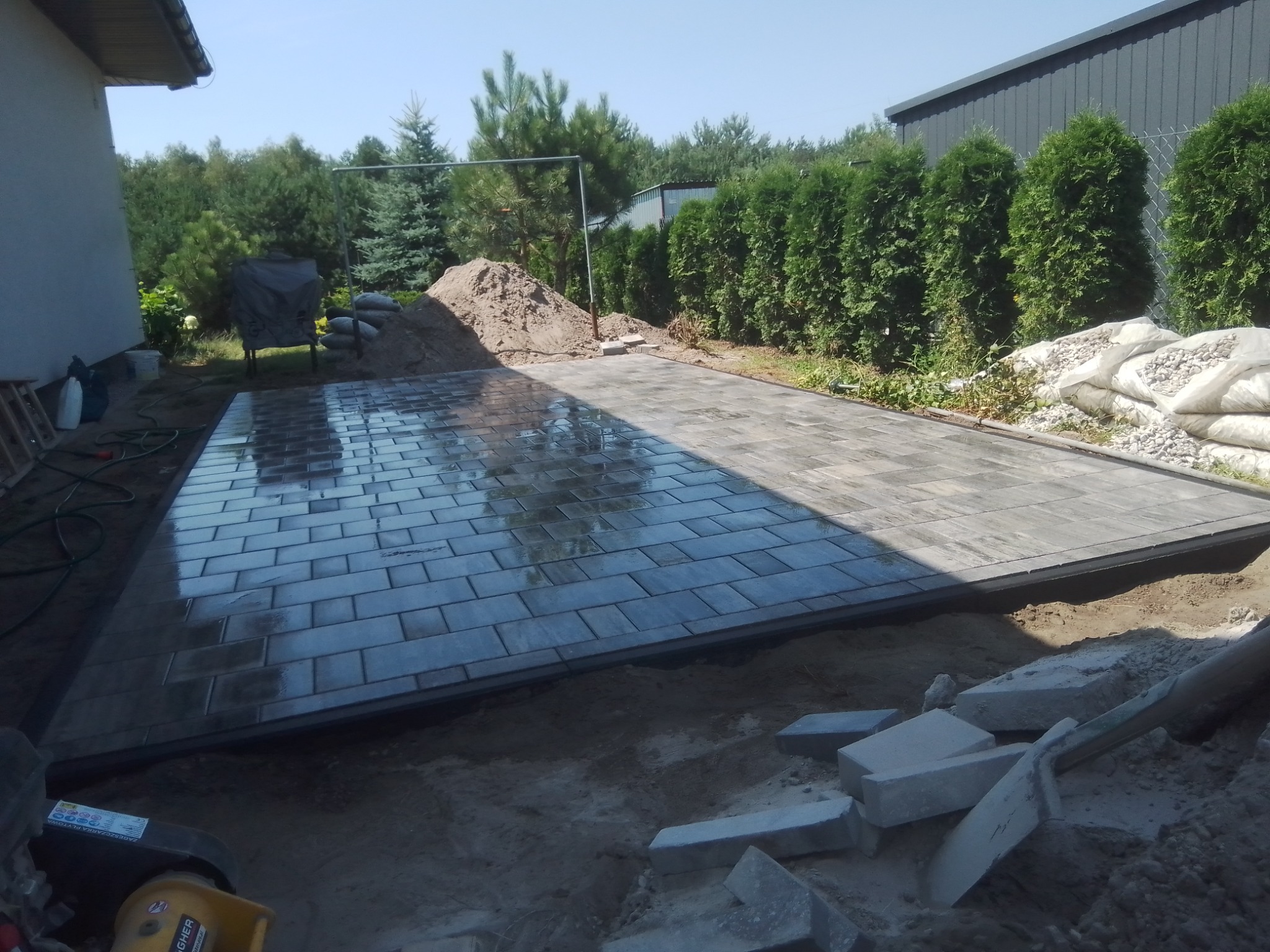 Układanie kostki brukowej w trakcie prac: widoczna część ułożonej, mokrej kostki, hałda piasku, worki z kruszywem i narzędzia na placu budowy w słoneczny dzień.