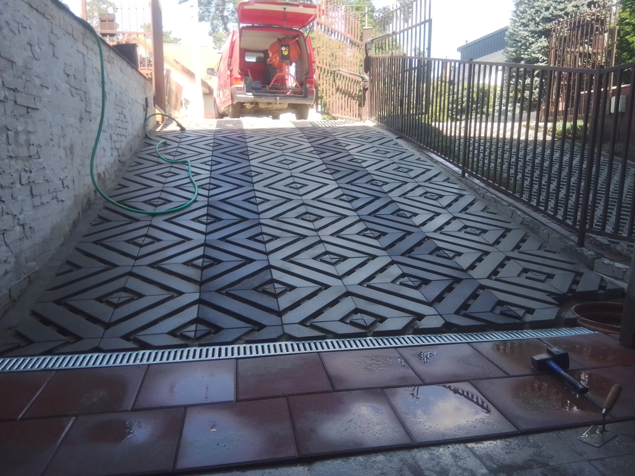 Ułożona nawierzchnia z szarej kostki o geometrycznym wzorze w kształcie rombów, oddzielona od brązowej kostki drenażem liniowym; w tle czerwony samochód dostawczy z otwartym bagażnikiem i narzędziami.