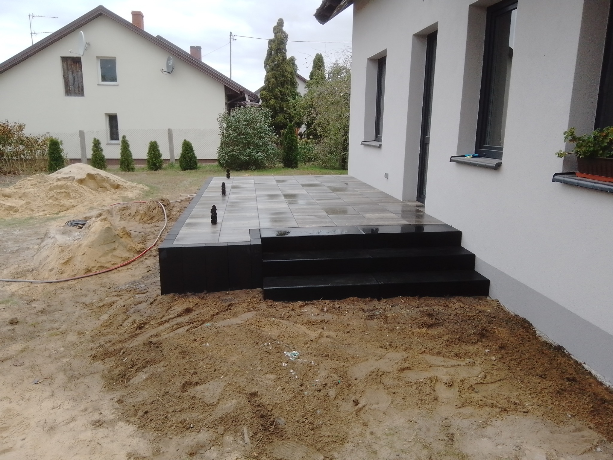 Nowo ułożony taras z szarych płyt z czarnym obramowaniem i schodami, widoczny piasek i ziemia dookoła, dom z jasną elewacją w tle.