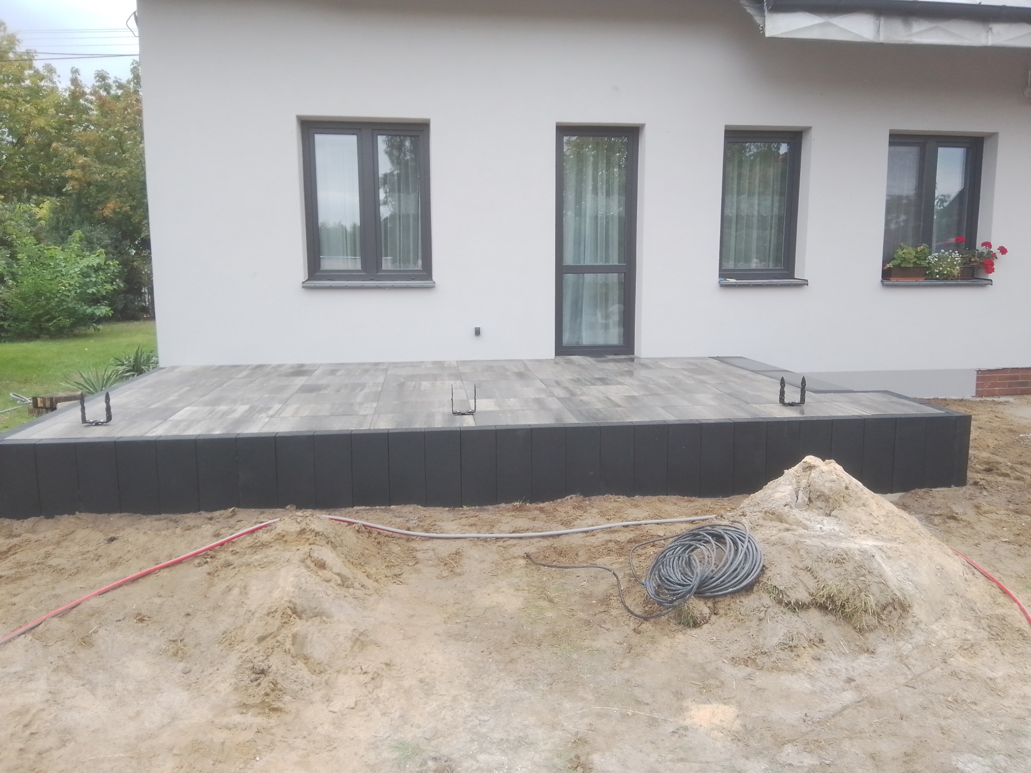Nowoczesny taras z szarej kostki brukowej, obrzeża z czarnych elementów, widok na dom z trzema oknami, w tle ogród, na pierwszym planie piasek i zwój kabla.
