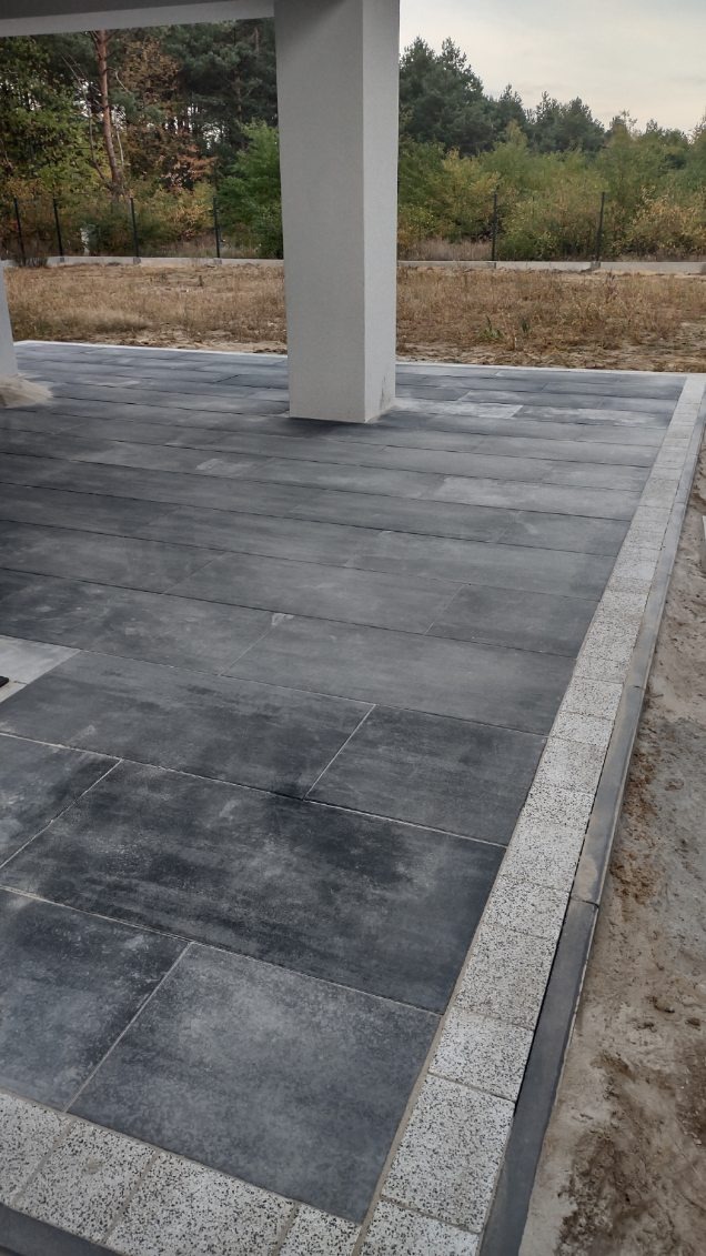 Nowoczesny taras z ciemnoszarej kostki brukowej o prostokątnym kształcie, obrzeża wykończone jasnoszarą kostką granitową, widoczny fragment kolumny nośnej budynku.