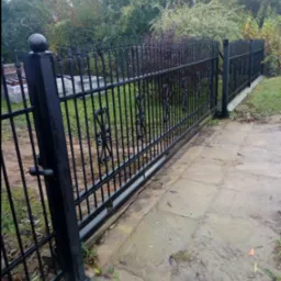 Czarna, metalowa brama przesuwna z ozdobnymi elementami w kształcie serc, prowadząca na posesję z widocznym ogrodem w tle.