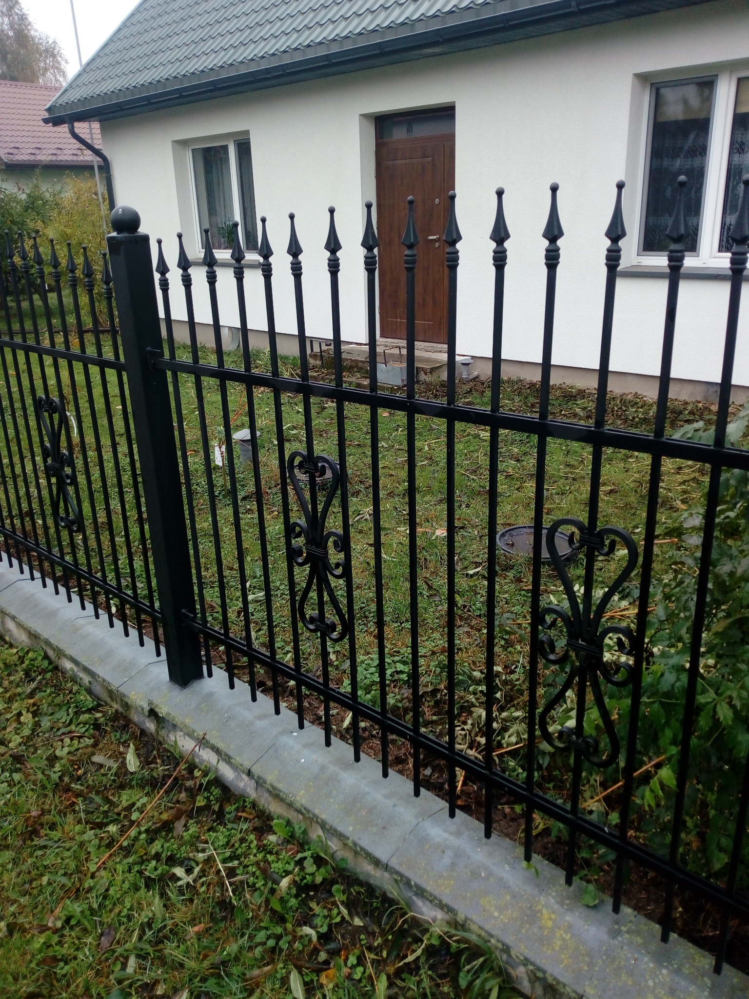 Czarny metalowy płot z ozdobnymi elementami w kształcie serc, otaczający trawnik przed białym domem z brązowymi drzwiami.