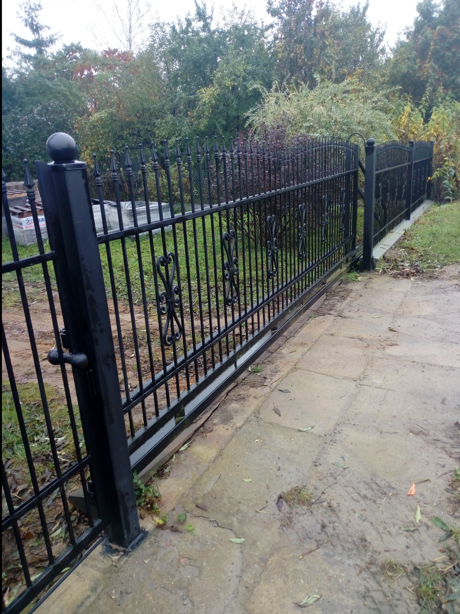 Czarna, metalowa brama przesuwna z ozdobnymi elementami w kształcie serc, prowadząca na posesję z widocznym ogrodem w tle.