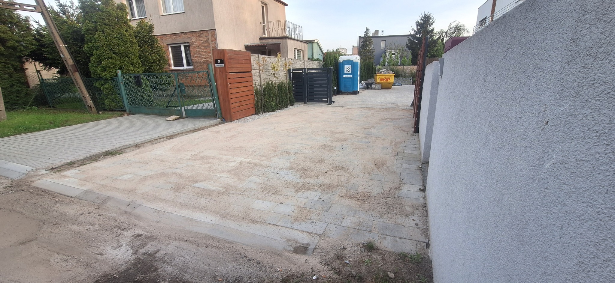 Układanie kostki brukowej na podjeździe do posesji, widoczna przenośna toaleta i kontener na gruz w tle.