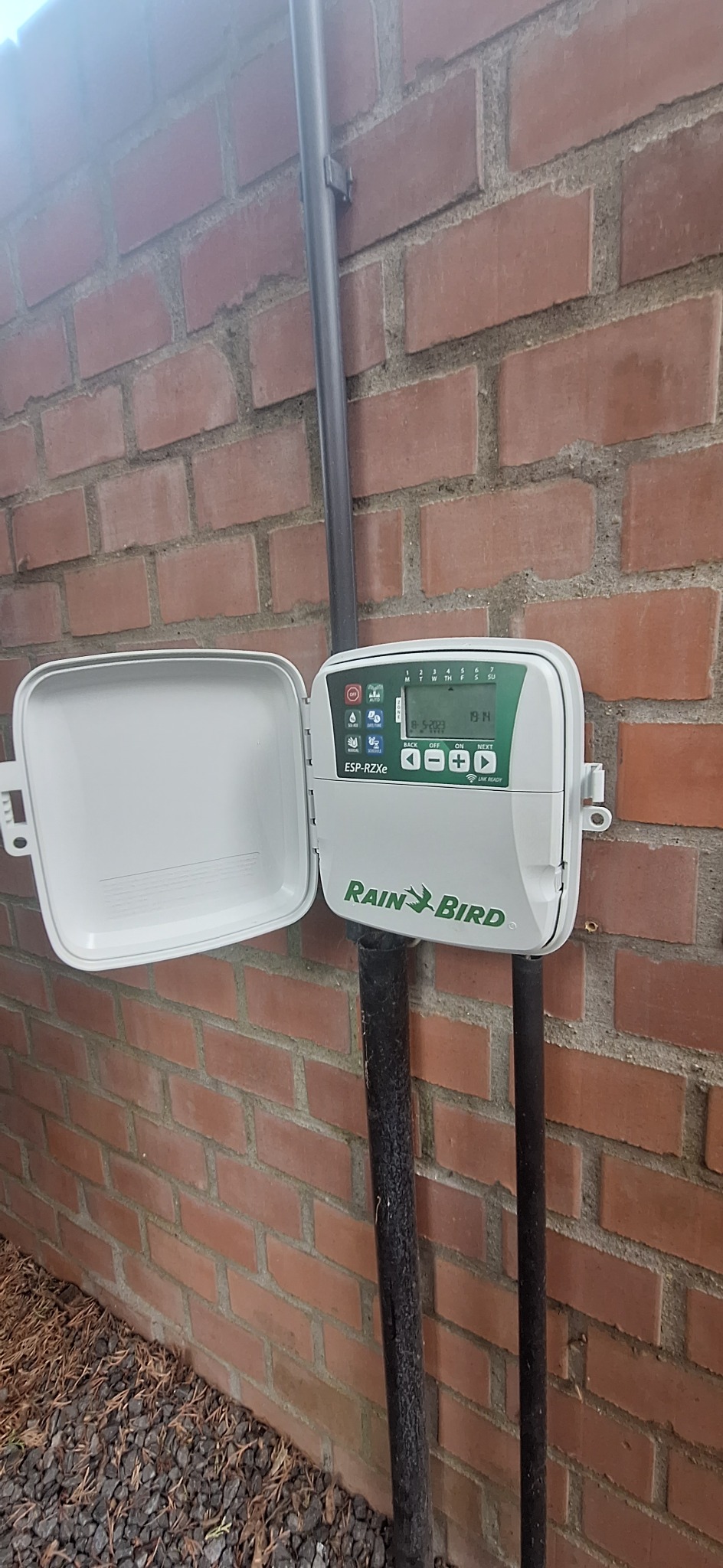 Sterownik nawadniania Rain Bird ESP-RZXe z otwartą klapką, zamontowany na ceglanej ścianie budynku, widoczne rury doprowadzające wodę i małe kamienie na ziemi