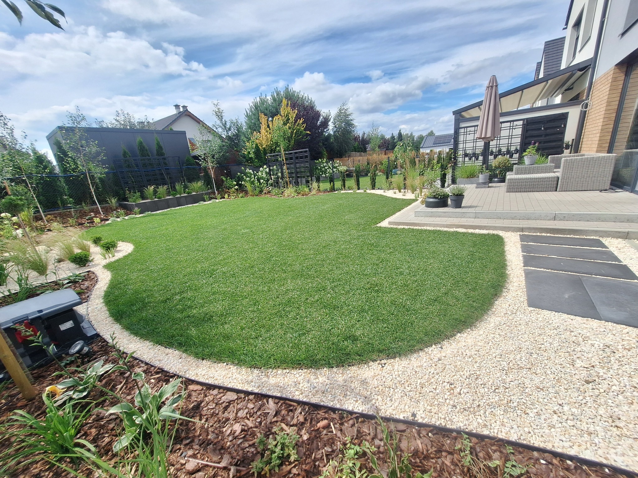 Zadbany trawnik z obrzeżem z jasnych kamieni i korą drzewną, otoczony roślinnością, z widokiem na nowoczesny dom z tarasem i pergolą w tle.