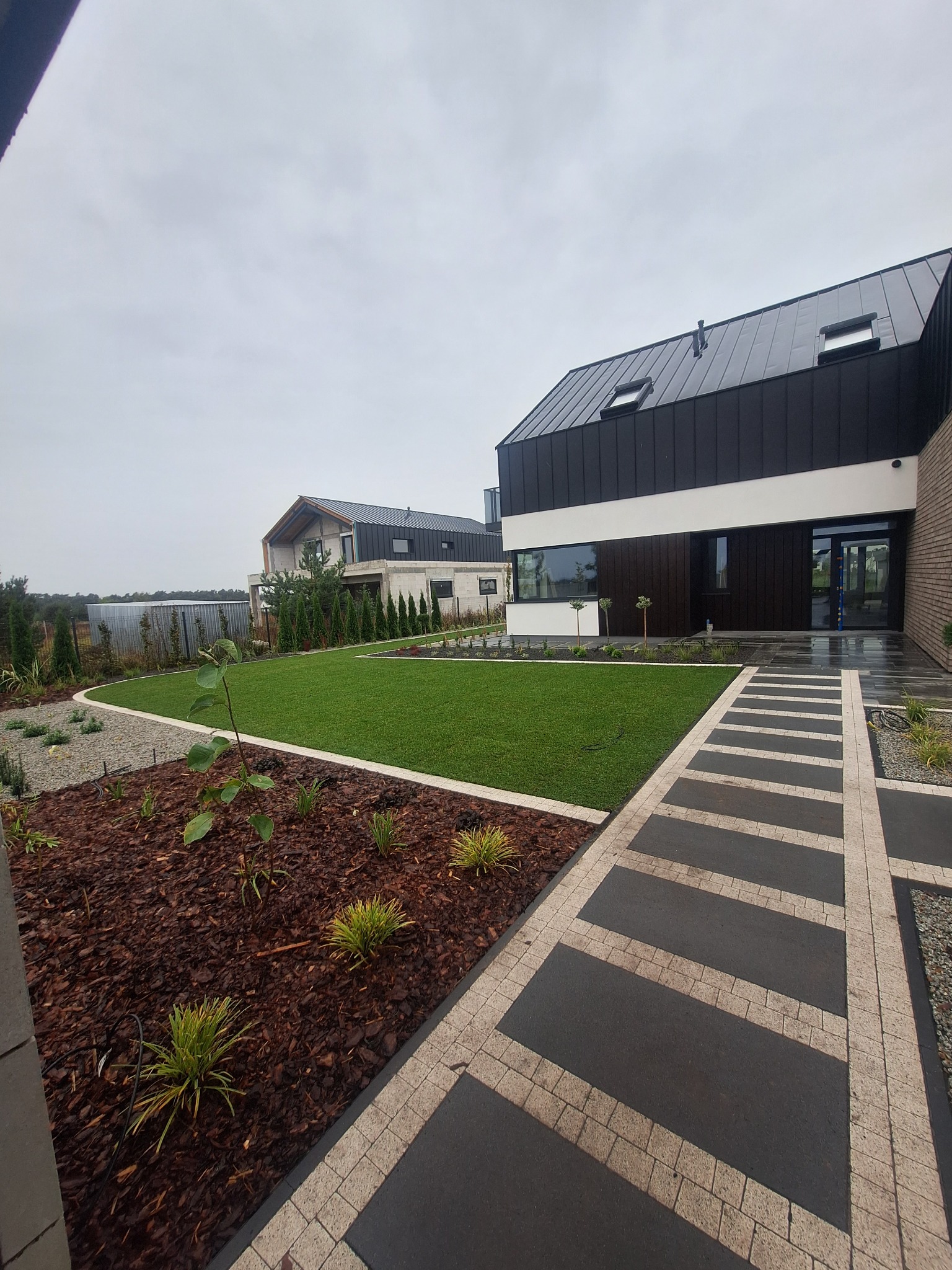 Nowoczesny ogród z geometrycznym trawnikiem, ścieżką z prostokątnych płyt i korą drzewną, otaczający dom z ciemną elewacją i skośnym dachem.