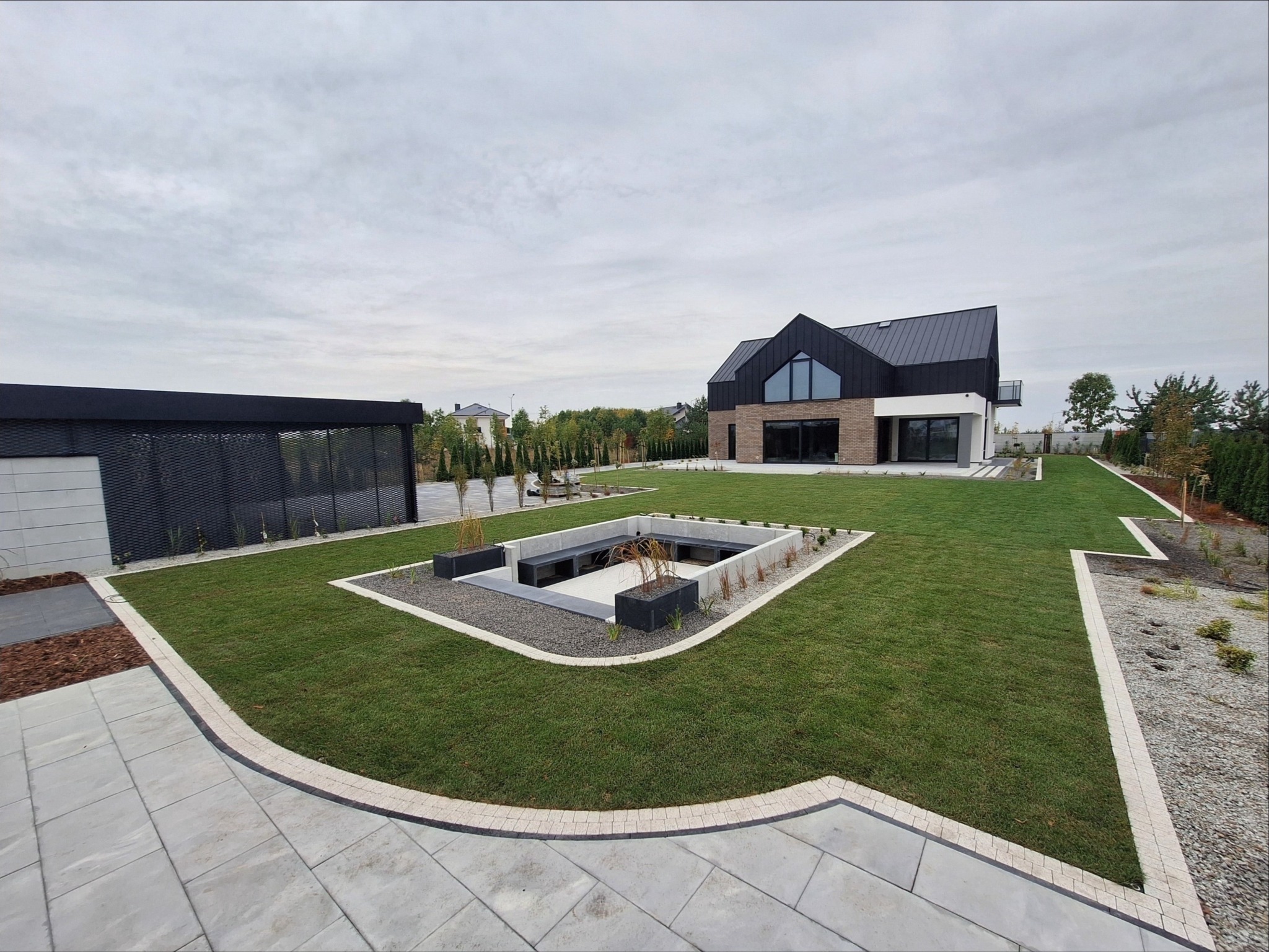 Nowoczesny ogród z równo przystrzyżonym trawnikiem, minimalistyczną strefą relaksu z betonowymi elementami i ozdobnymi trawami, oraz ścieżką z jasnych płyt prowadzącą do domu o nowoczesnej...