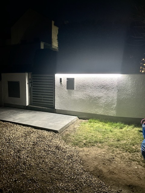 Oświetlona ściana z bramą i fragmentem trawnika w nocy, widoczne drobne kamienie na ziemi.