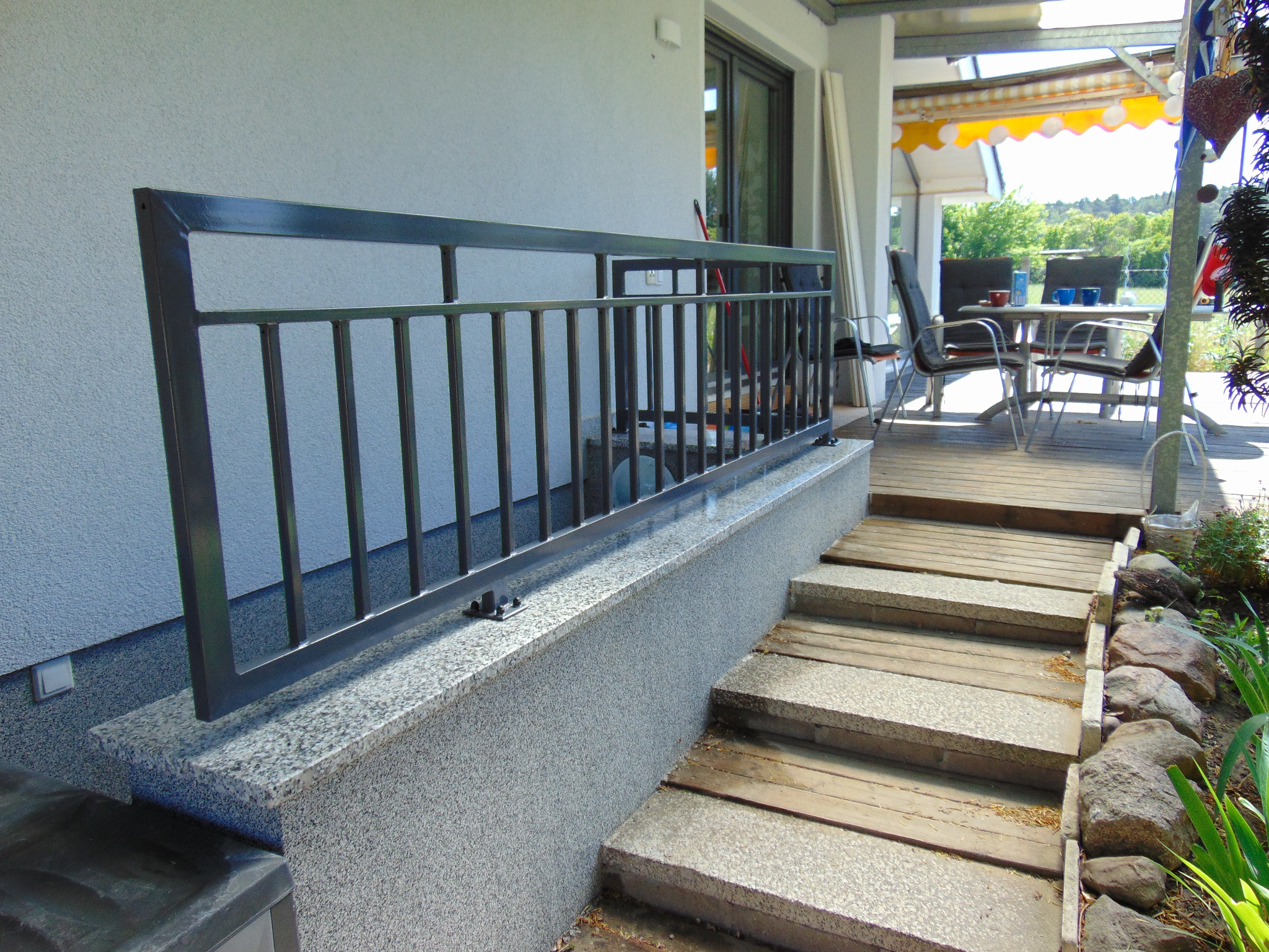 Metalowa balustrada w kolorze grafitowym zamontowana na niskim murku z szarego kamienia, prowadząca do drewnianego tarasu z meblami ogrodowymi i żółtą markizą.