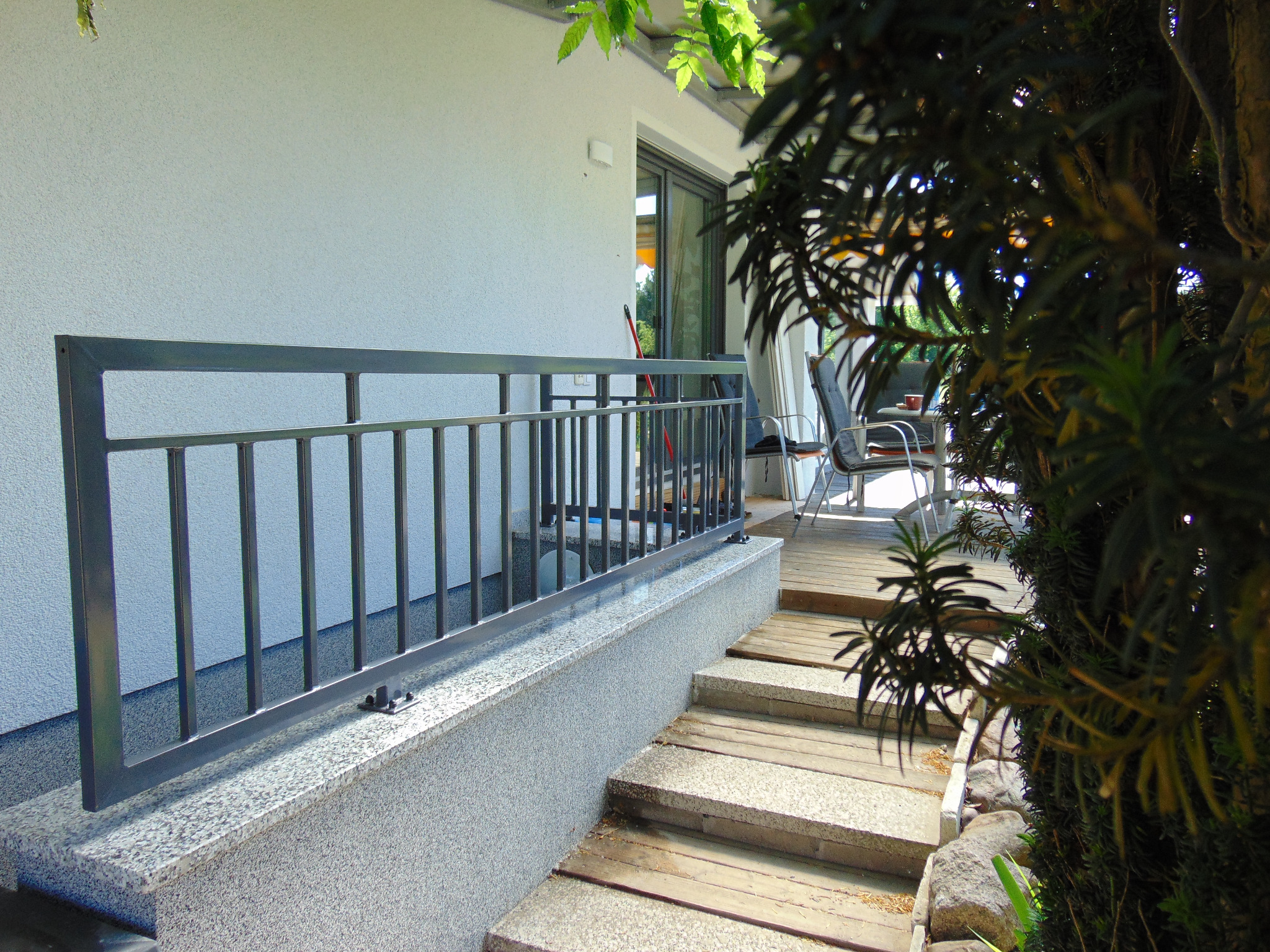 Metalowa balustrada w kolorze grafitowym zamontowana na podeście z lastryko, prowadząca do drewnianego tarasu z widocznymi schodami i meblami ogrodowymi w tle.