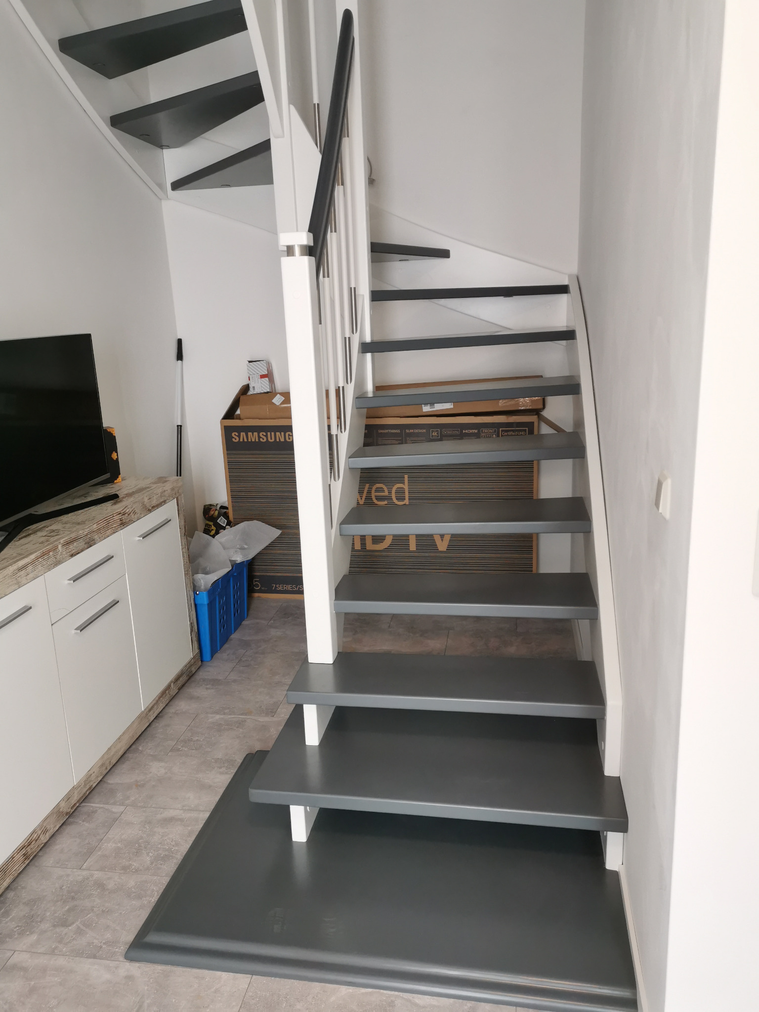 Wewnętrzne schody z drewnianymi stopniami pomalowanymi na szaro, białe barierki i czarny poręcz, widoczne z dołu, w tle szafka i kartony.