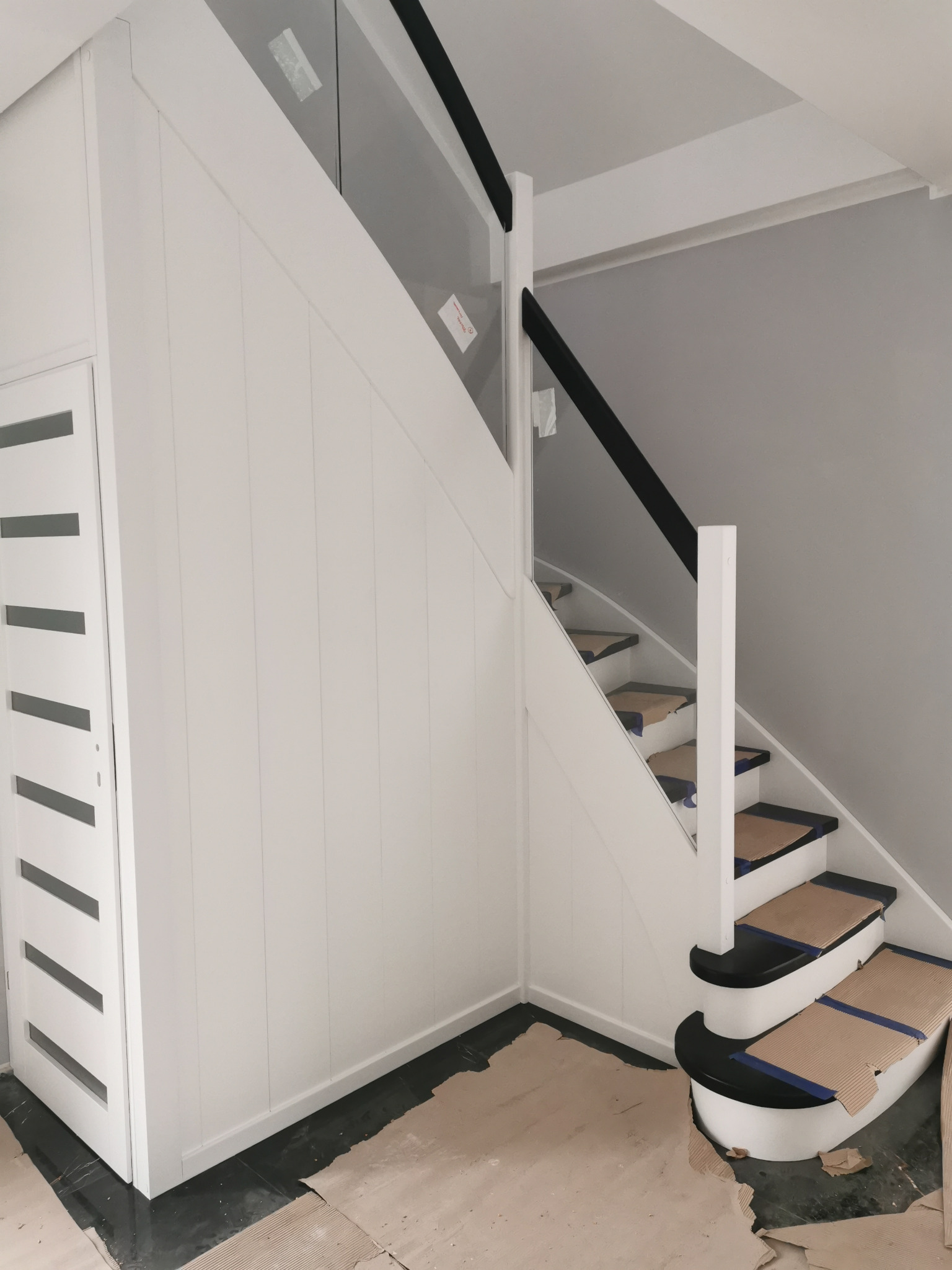 Białe schody z czarnymi akcentami i szklaną balustradą, zabezpieczone kartonem na stopniach podczas prac wykończeniowych.
