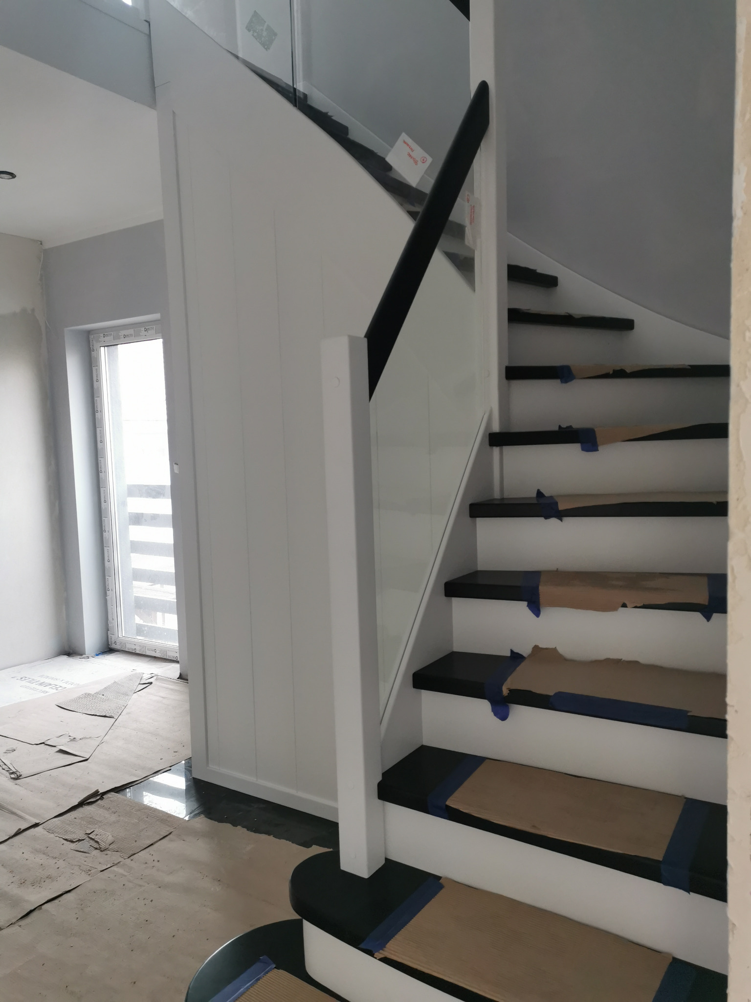 Wewnętrzne schody z czarnymi stopniami zabezpieczonymi kartonem i niebieską taśmą, białe podstopnice i biała balustrada z elementami szklanymi, czarna poręcz, widok w trakcie montażu lub wykończenia.