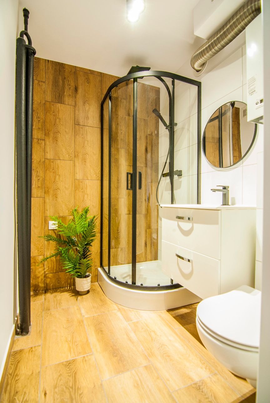 Nowoczesna łazienka z kabiną prysznicową z czarnymi ramami, drewnopodobnymi płytkami na ścianie i podłodze, białą szafką umywalkową i okrągłym lustrem. Na podłodze stoi doniczka z zieloną rośliną.
