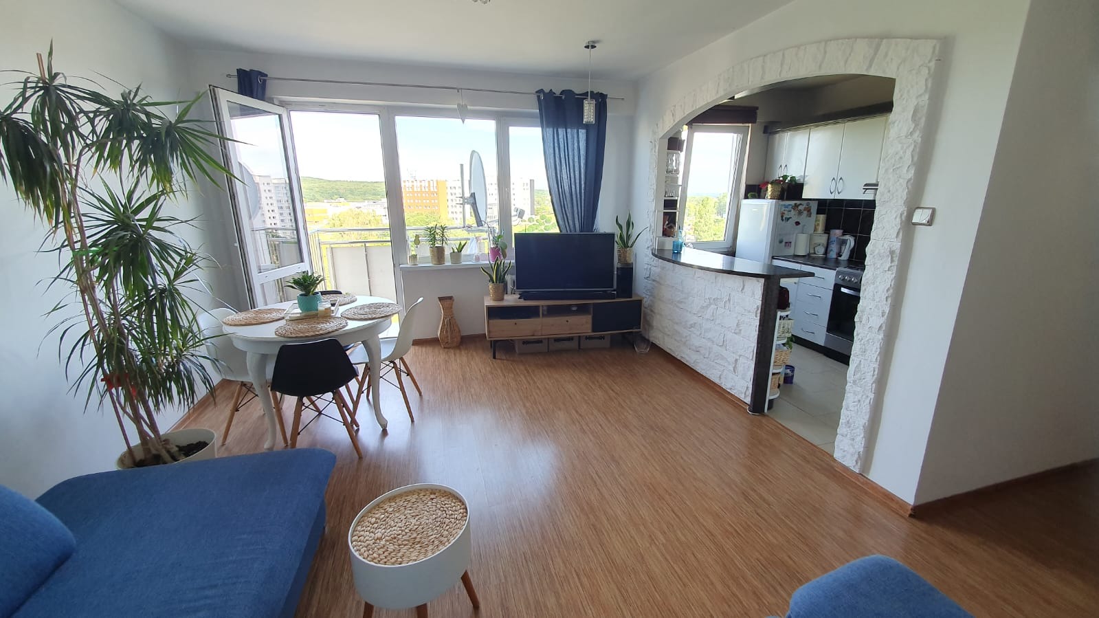 Jasne wnętrze mieszkania z drewnianą podłogą, okrągłym stołem jadalnym z trzema krzesłami i jednym czarnym, telewizorem na szafce RTV, roślinami doniczkowymi przy balkonie i otwartą kuchnią...