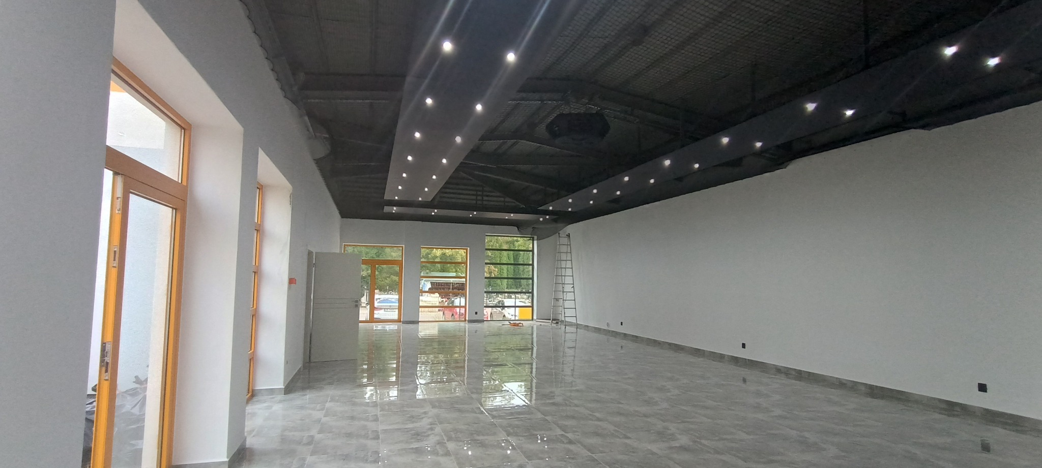 Wnętrze przestronnego, pustego lokalu z szarą, błyszczącą podłogą, białymi ścianami, oknami z drewnianymi ramami i czarnym sufitem z punktowym oświetleniem; widoczna drabina oparta o ścianę.