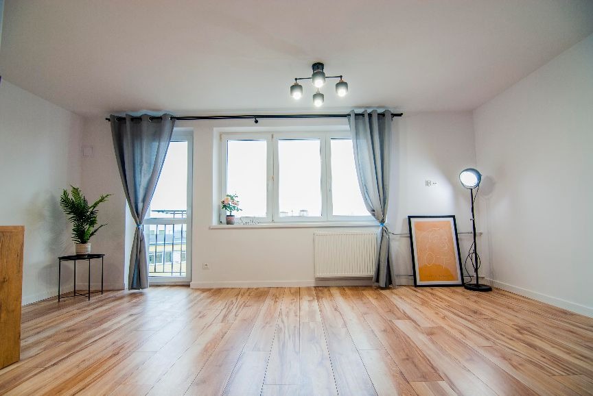 Jasne, przestronne wnętrze pokoju z drewnianą podłogą, dużym oknem z szarymi zasłonami, rośliną w doniczce na stoliku, grzejnikiem pod oknem, nowoczesną lampą stojącą i plakatem w czarnej ramie...