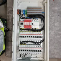 Otwarta, biała skrzynka rozdzielcza z zainstalowanymi bezpiecznikami, ogranicznikami przepięć (czerwone) i listwami zaciskowymi. Widoczne przewody w czarnej izolacji. Skrzynka zamontowana na szarej...