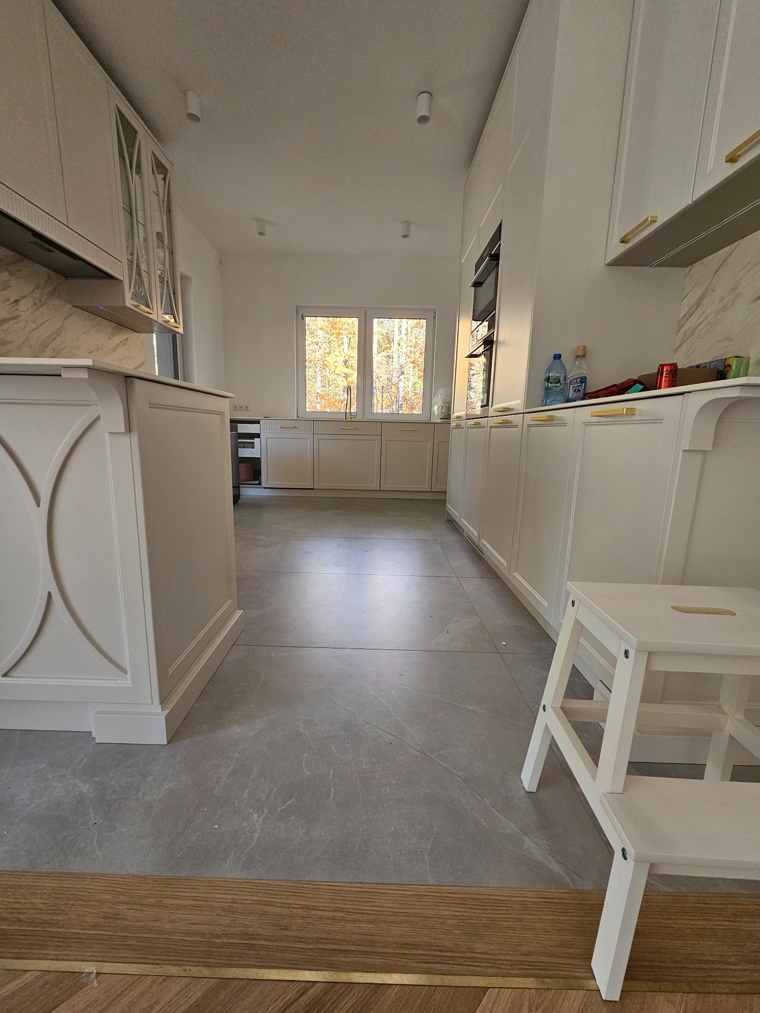 Nowoczesna, jasna kuchnia z białymi szafkami z złotymi uchwytami, szarymi płytkami podłogowymi imitującymi marmur i drewnianym progiem, widok z niskiej perspektywy.