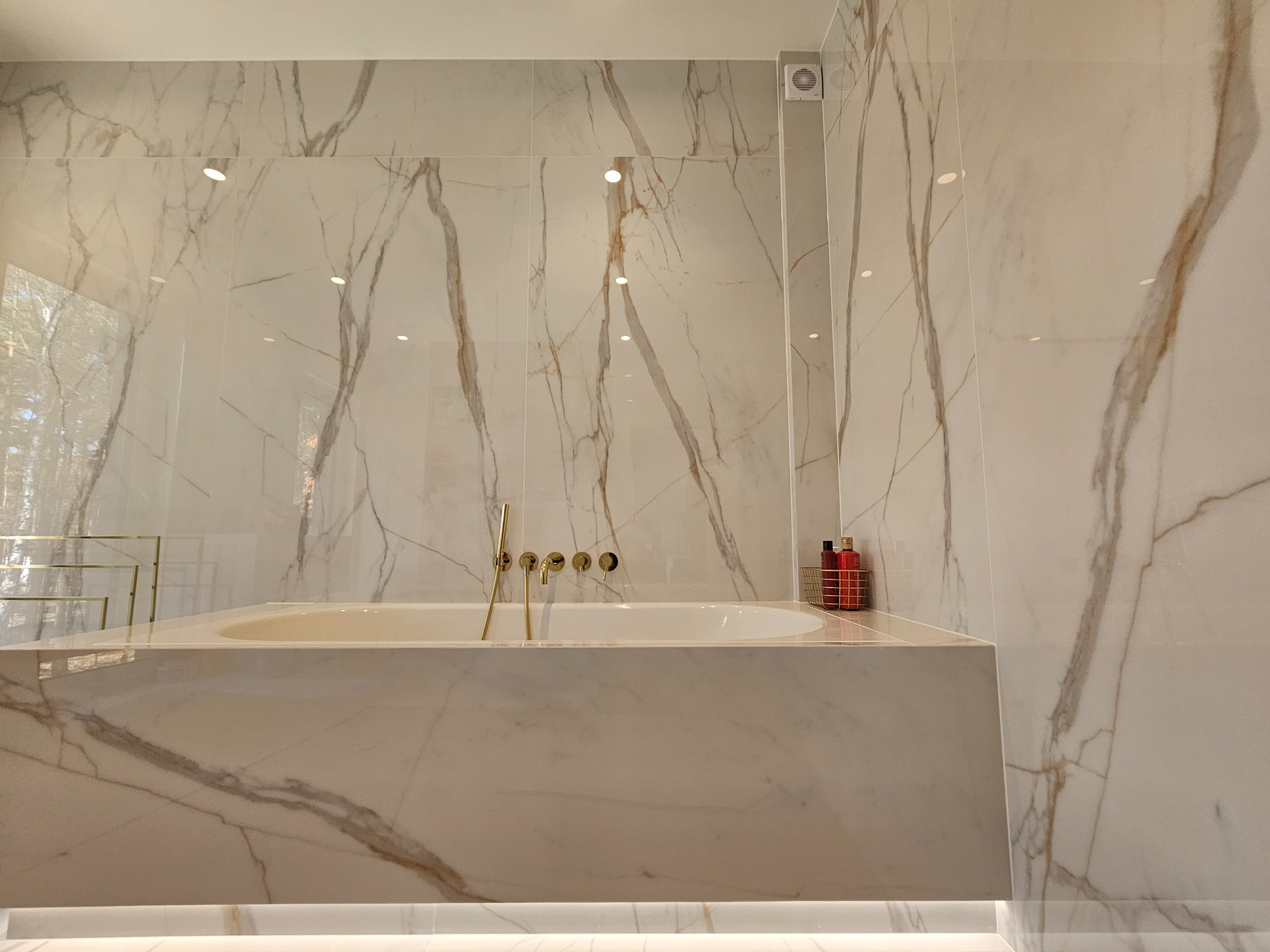 Luksusowa łazienka z wanną obudowaną marmurem Calacatta, złota armatura ścienna i podświetlenie LED u dołu obudowy wanny.