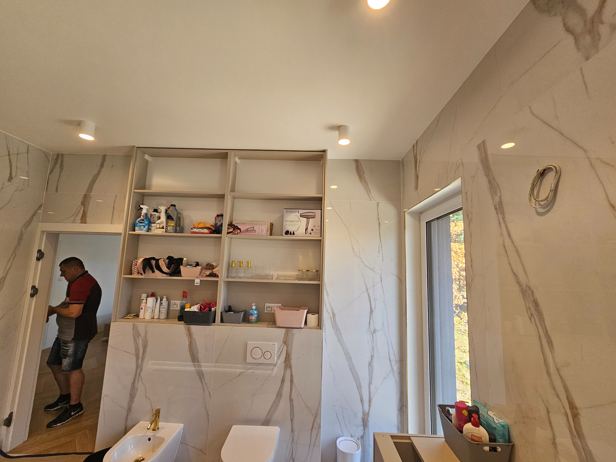 Wnętrze łazienki z marmurowymi płytkami, widoczna zabudowa regałowa z półkami wypełnionymi kosmetykami i akcesoriami, oraz częściowo zamontowana armatura: bidet i toaleta. Pracownik w tle...