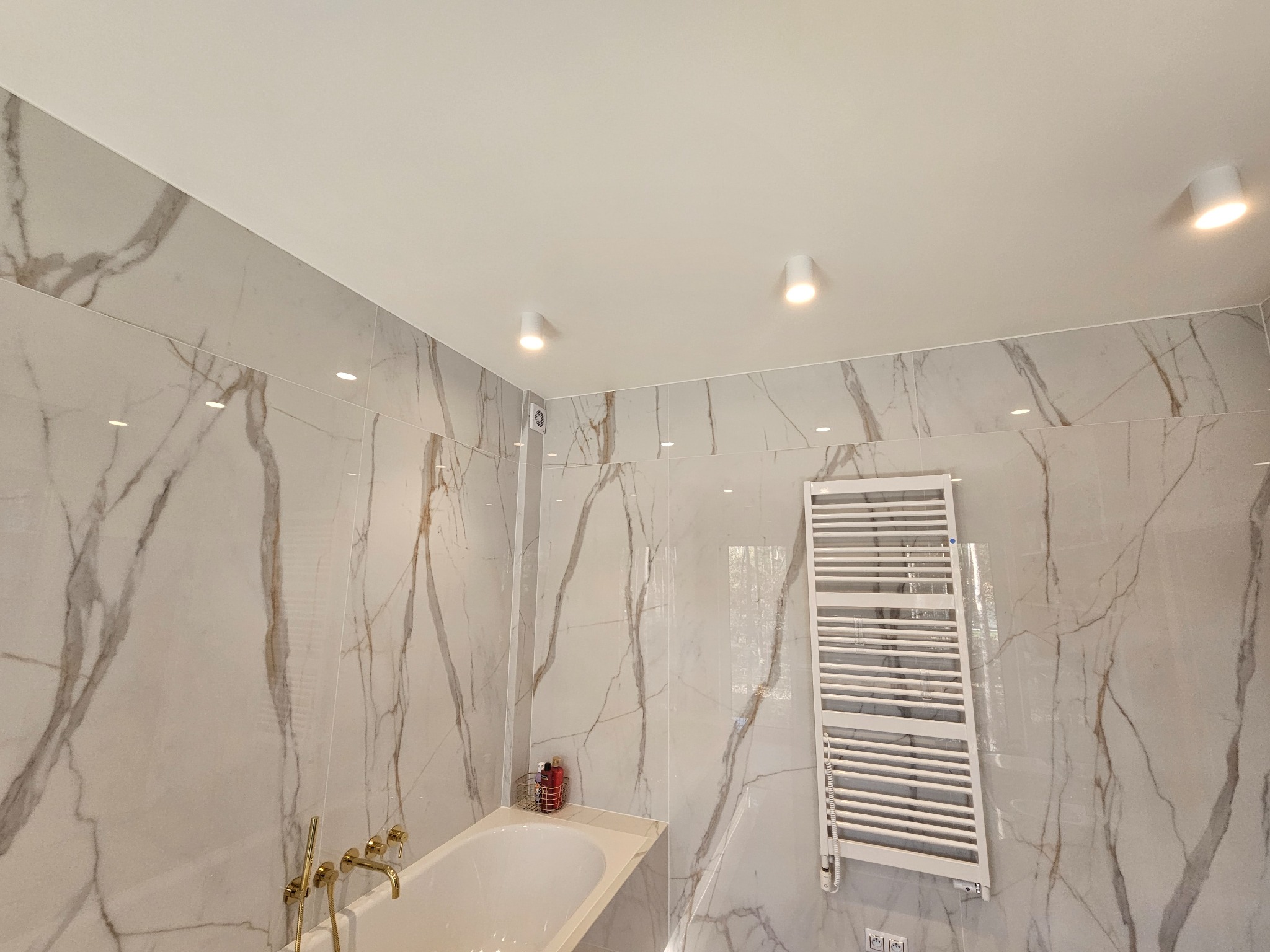 Elegancka łazienka wykończona płytkami imitującymi marmur z złotą armaturą, grzejnikiem drabinkowym i oświetleniem punktowym.