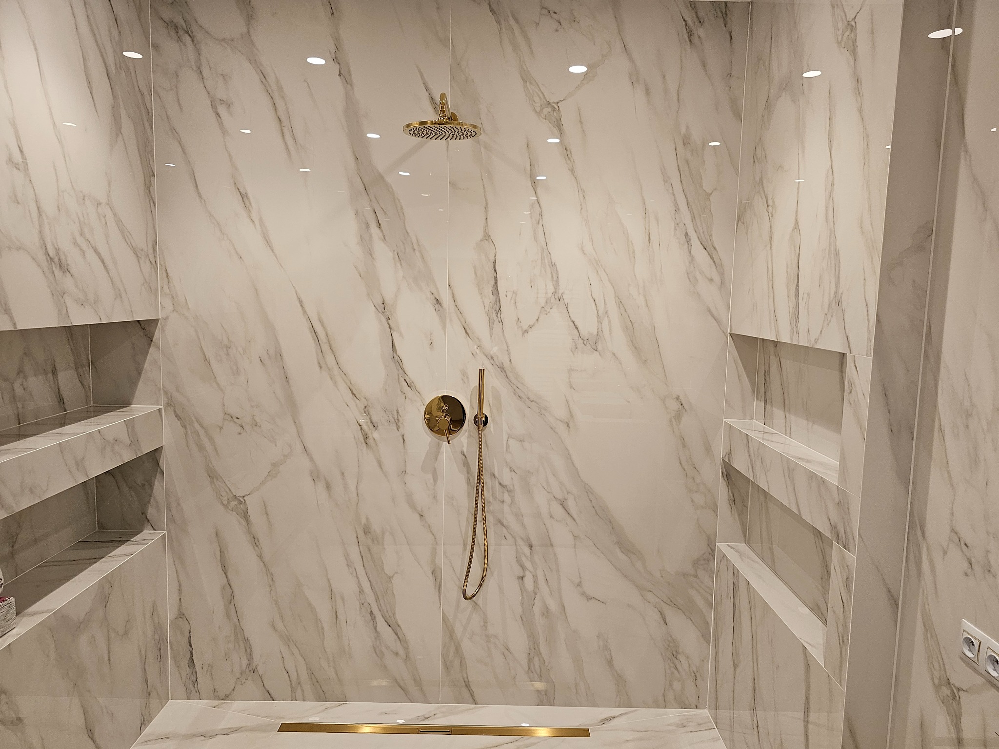 Kabina prysznicowa z marmurowymi płytkami, złotą armaturą i wnękami półkowymi po bokach, wykończona listwą odpływową w kolorze złota.