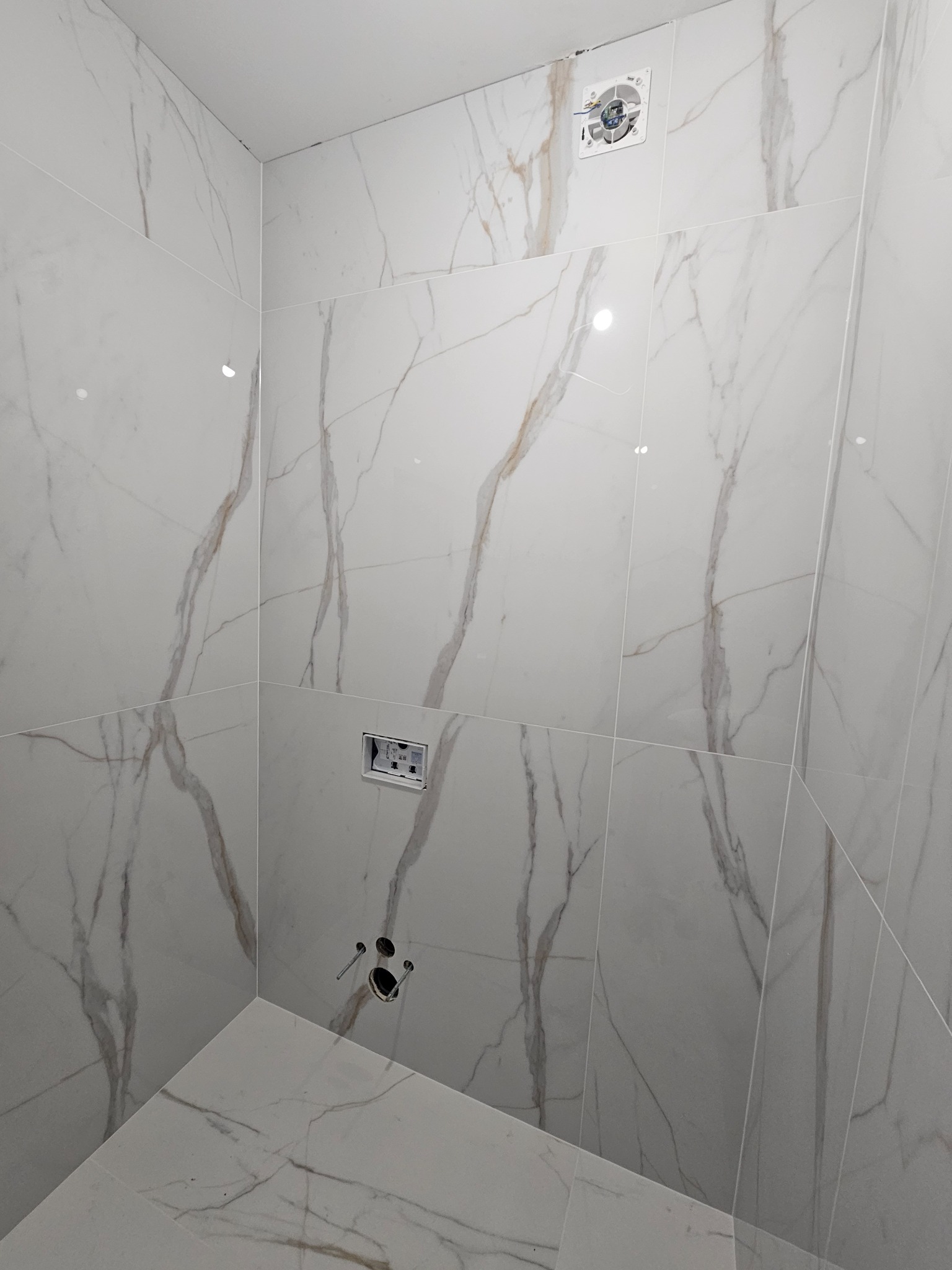 Ściany i podłoga w łazience wyłożone białymi płytkami imitującymi marmur z widocznymi przyłączami do armatury i wentylatorem sufitowym.