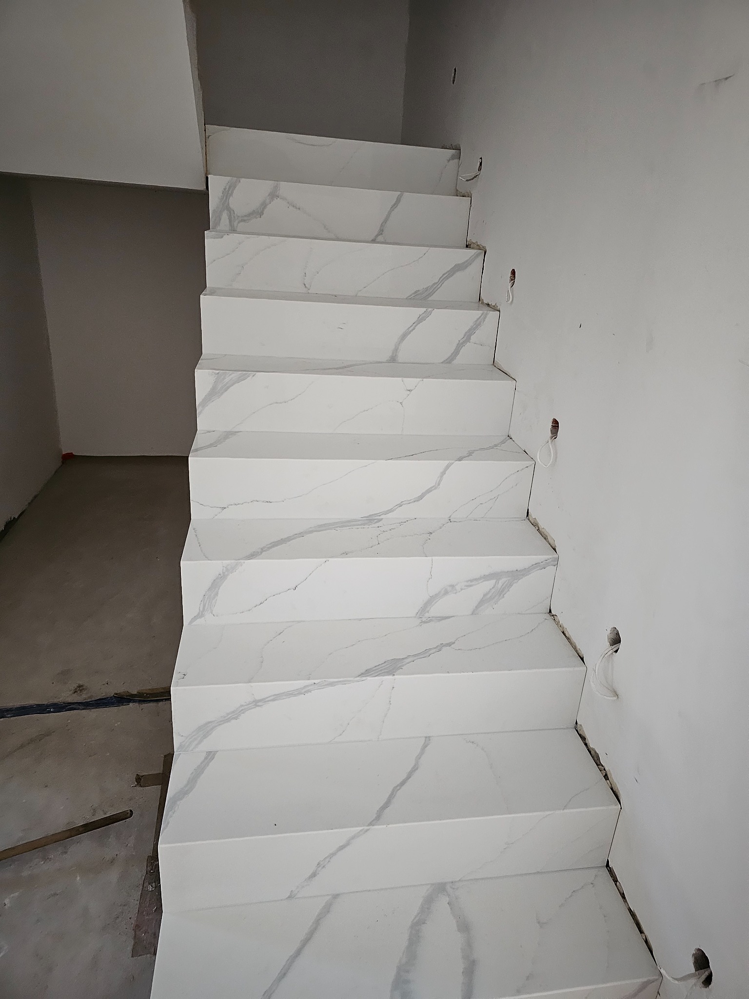 Schody wyłożone białym gresem imitującym marmur w trakcie montażu, widoczne niedoskonałości i przygotowane otwory na kable w ścianie, Warszawa.