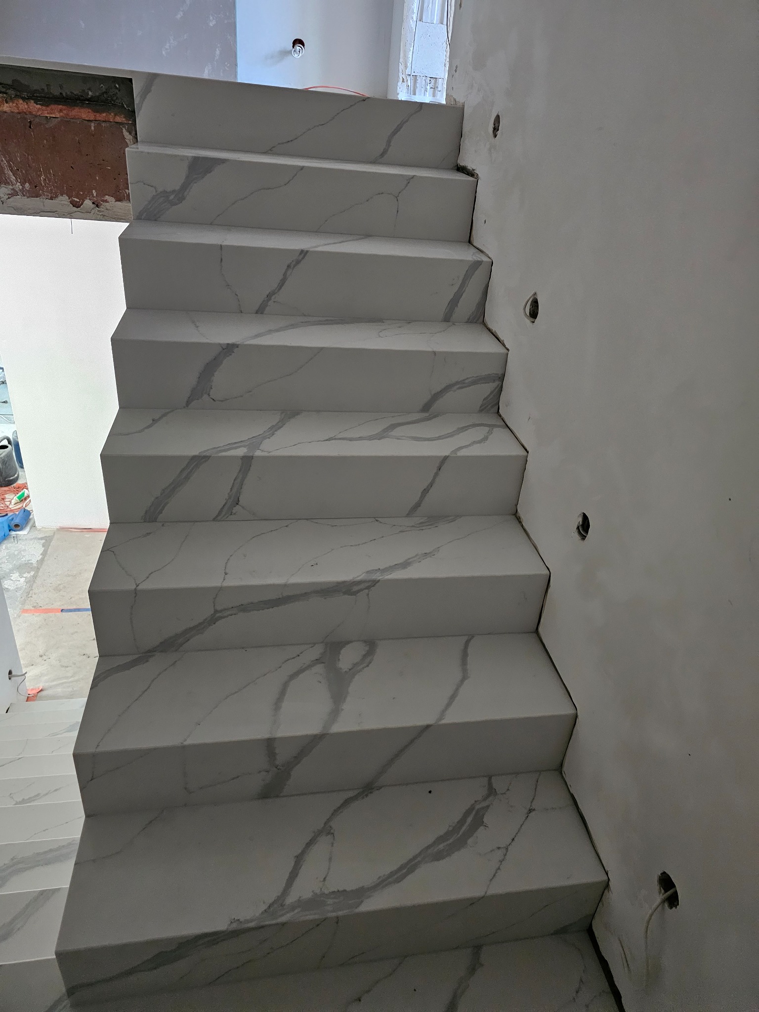 Schody wykończone białym gresem imitującym marmur, widoczne niedokończone ściany i instalacja elektryczna.