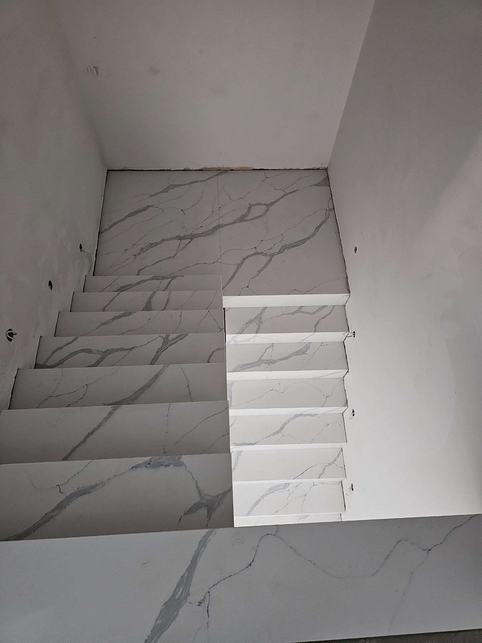 Schody wyłożone białymi płytami z szarym żyłkowaniem, widok z góry, surowe ściany w tle.
