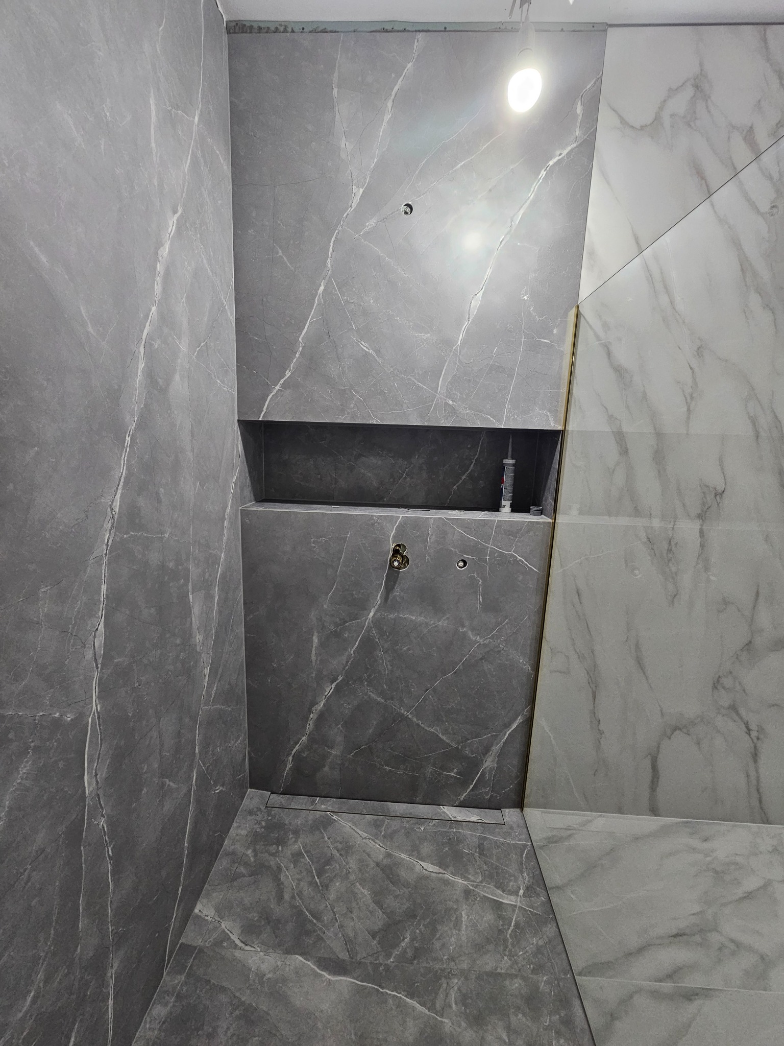 Szara łazienka z marmurowymi płytkami, wnęka z silikonem, widoczne przyłącza wodne.