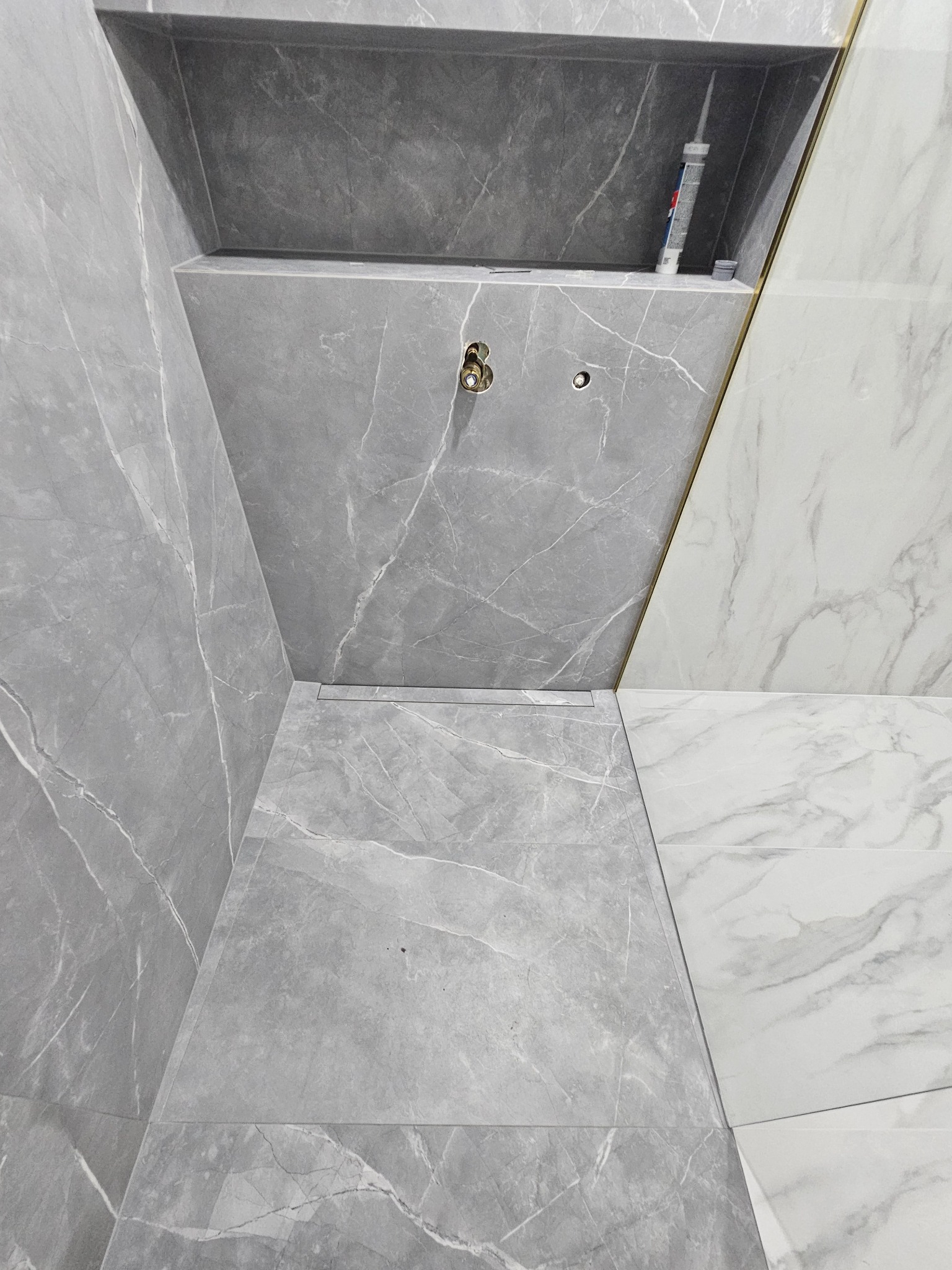 Nowoczesny prysznic walk-in wyłożony szarymi płytkami imitującymi marmur, z odpływem liniowym i wnęką na kosmetyki, w której widoczna jest tuba silikonu.