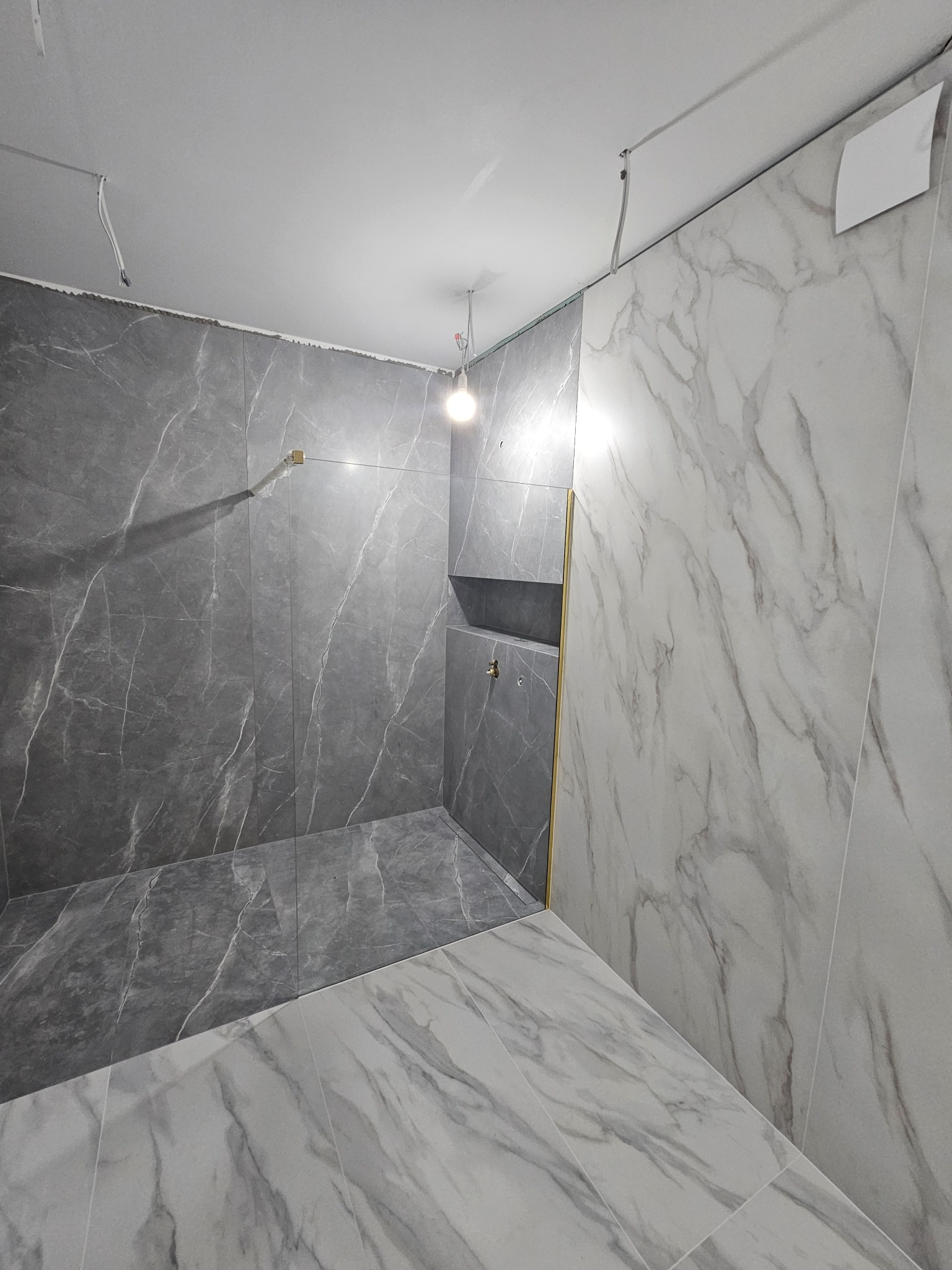 Niedokończona łazienka z szarymi i białymi płytkami imitującymi marmur, widoczne wystające kable i brakujące fugi na suficie, wnęka prysznicowa z złotym wykończeniem.