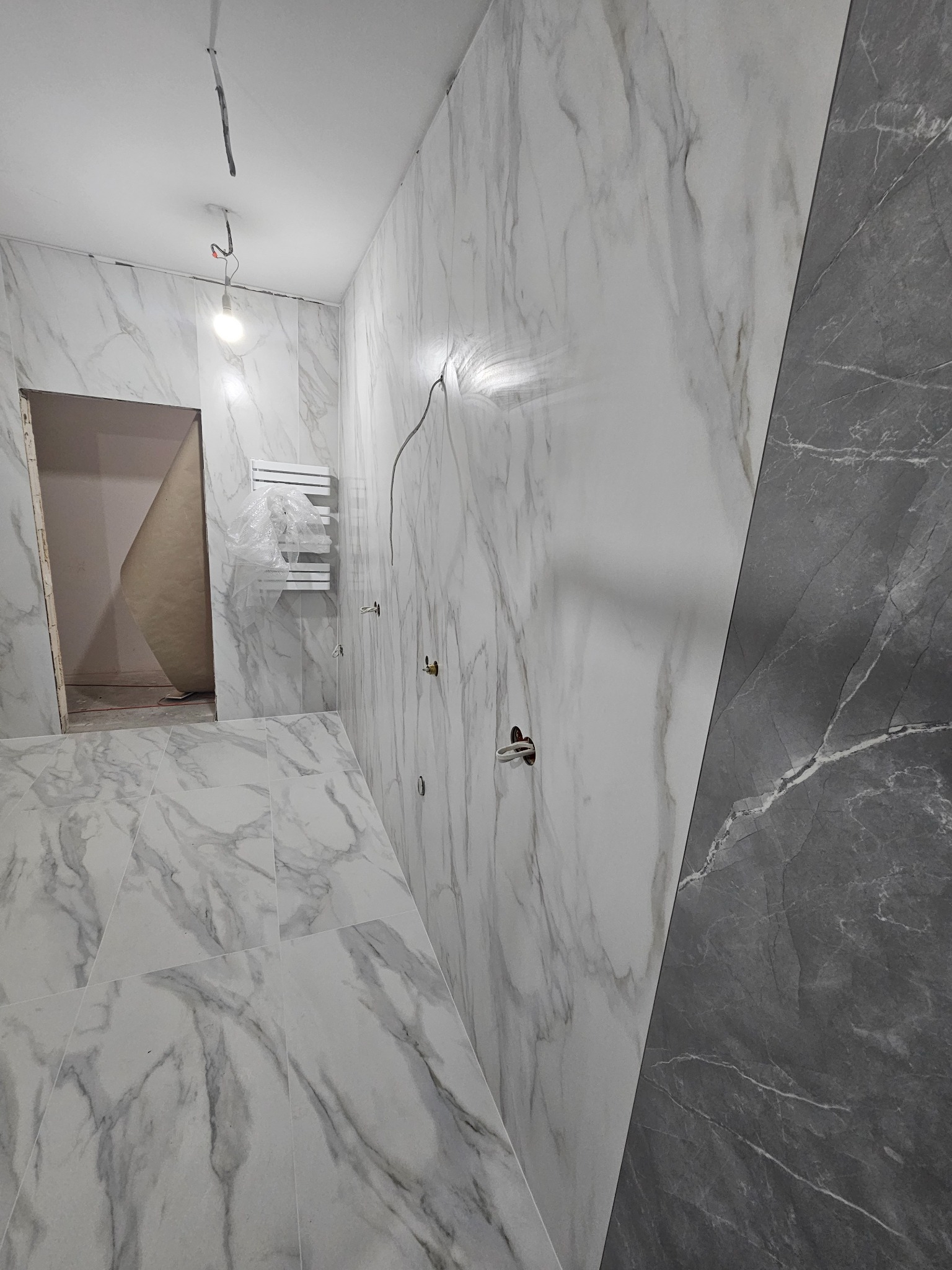 Łazienka w trakcie remontu: białe i szare płytki marmurowe na ścianach i podłodze, widoczne wyprowadzenia elektryczne, grzejnik drabinkowy w folii ochronnej, surowy strop i wisząca żarówka.