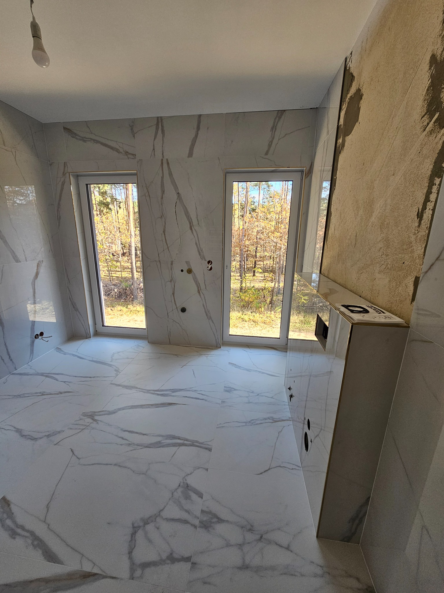 Wnętrze łazienki w trakcie remontu, z marmurowymi płytkami na podłodze i ścianach, widoczne dwa okna z widokiem na las, niedokończona ściana z widocznymi śladami zaprawy, oraz wisząca żarówka bez...