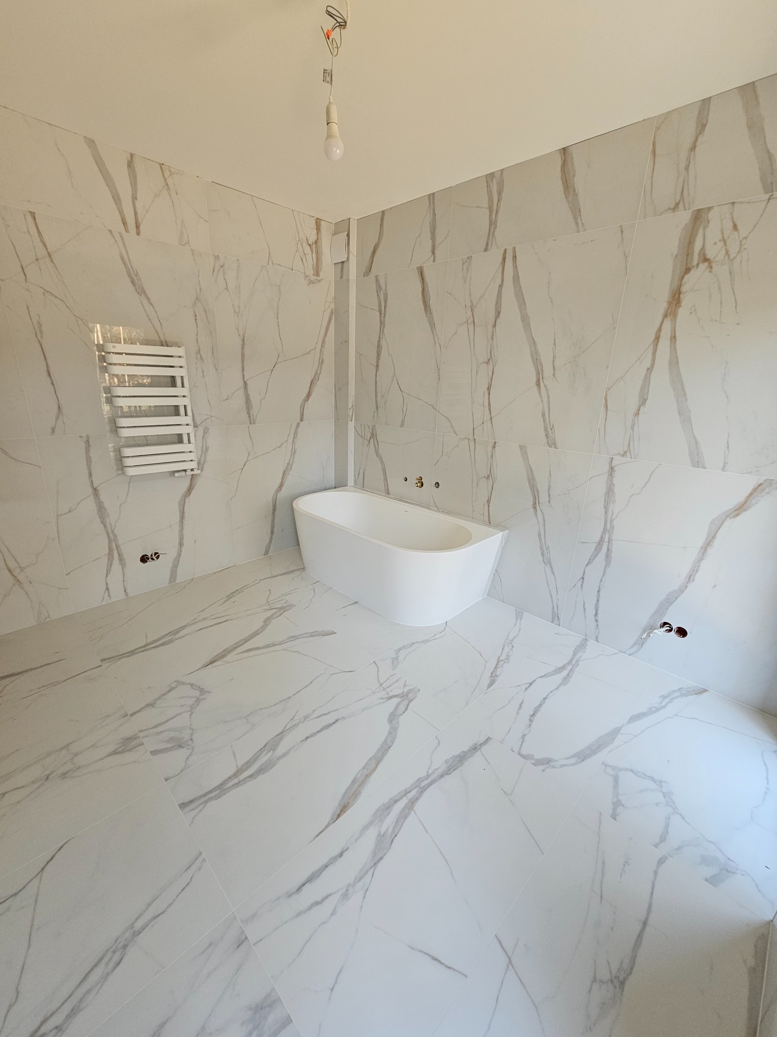 Nowa, jasna łazienka z białymi płytkami imitującymi marmur na ścianach i podłodze, z wolnostojącą wanną i grzejnikiem drabinkowym, widoczne przygotowane punkty instalacji wodnej, surowa żarówka...