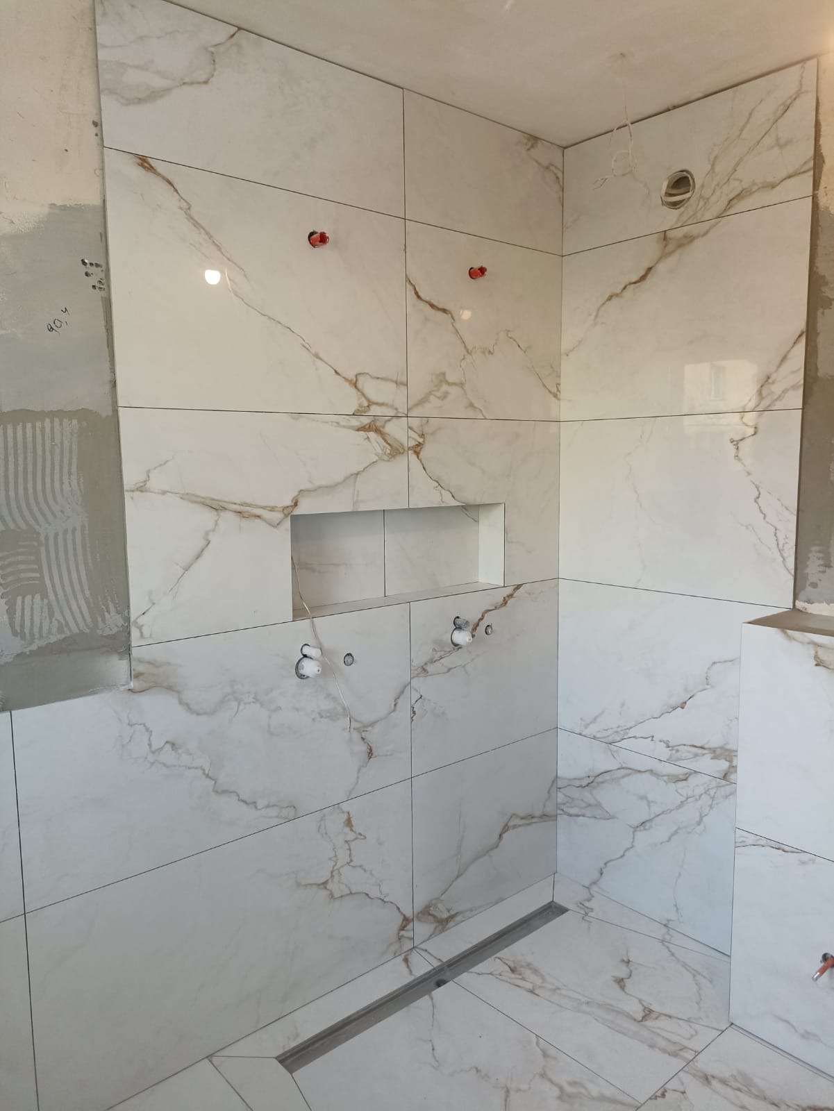 Łazienka w trakcie wykańczania: białe płytki imitujące marmur z subtelnymi złotymi żyłkami, wbudowana półka w ścianie, odpływ liniowy w podłodze, widoczne wyprowadzenia instalacji wodnej...