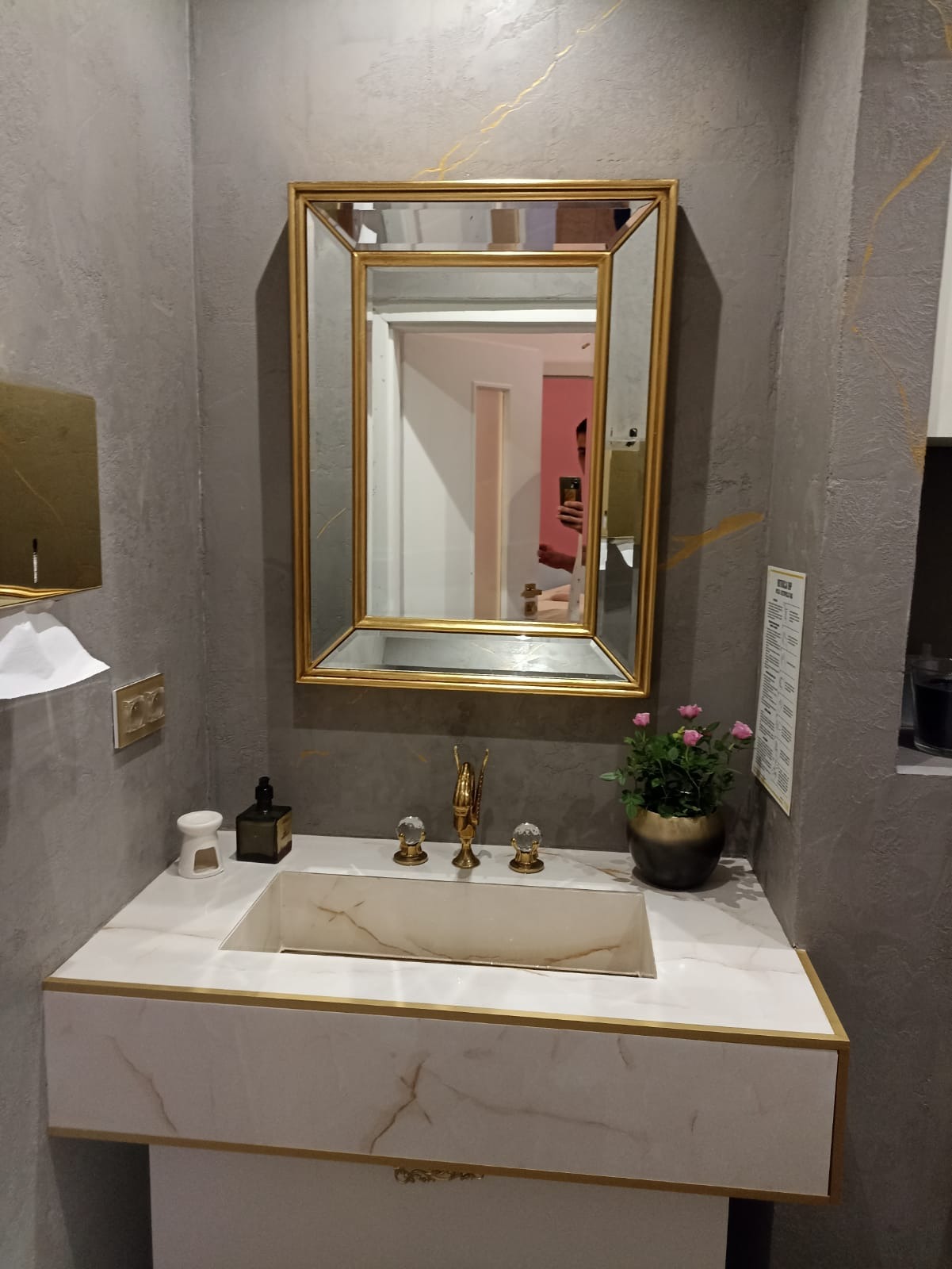 Nowoczesna łazienka z umywalką z marmurowym wzorem, złotą armaturą i lustrem w złotej ramie, na tle szarej ściany z dekoracyjnymi złotymi żyłkami.