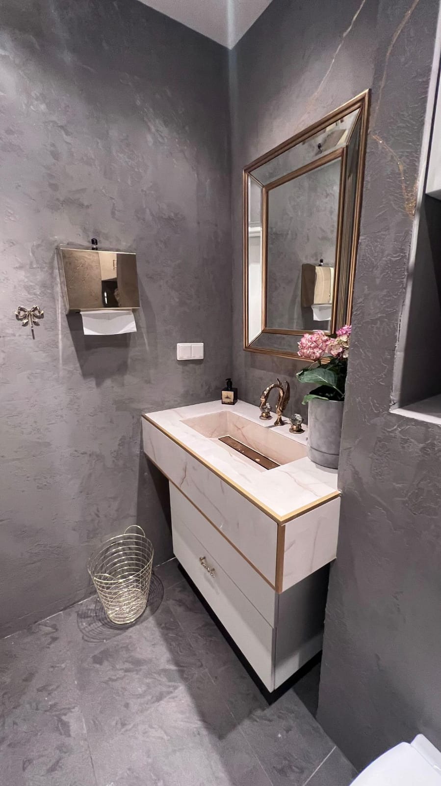 Elegancka łazienka w odcieniach szarości z marmurową umywalką, złotymi akcentami i lustrem w złotej ramie, uzupełniona o dekoracyjny kosz na śmieci i dozownik do papieru w kolorze złota.