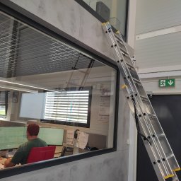 Clinox - Profesjonalne pranie kanap i tapicerki meblowej Warszawa - Aluminiowa drabina oparta o ścianę z oknem w biurze; widoczny pracownik przy biurku,  sufit z kratką, lampy liniowe i znak ewakuacyjny.
