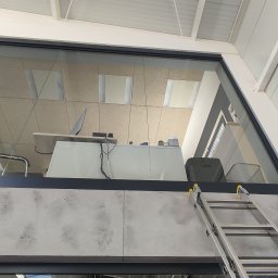 Clinox - Profesjonalne pranie kanap i tapicerki meblowej Warszawa - Wnętrze biura widziane z dołu przez szklaną ścianę, z widoczną drabiną opartą o ścianę, sugerującą prace porządkowe lub konserwacyjne.